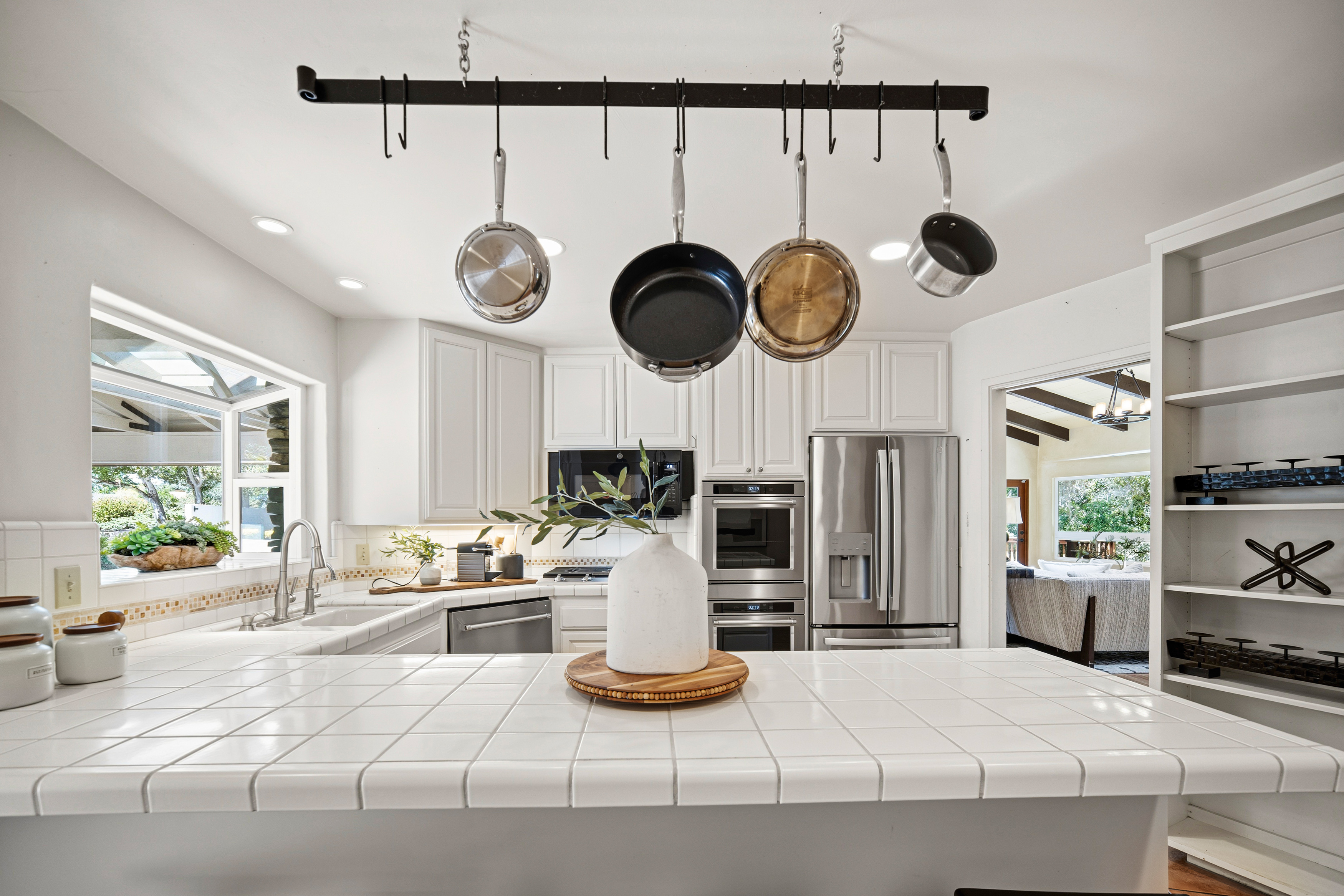 This bright, well-lit kitchen features white cabinetry, a tiled countertop island, and stainless steel appliances, including a double wall oven and a side-by-side refrigerator. A black metal pot rack hangs above the island, holding various cookware, while a decorative vase with greenery serves as a centerpiece. The perspective looks directly across the island toward the main cooking area, with an open doorway leading to an adjacent living space in the background.