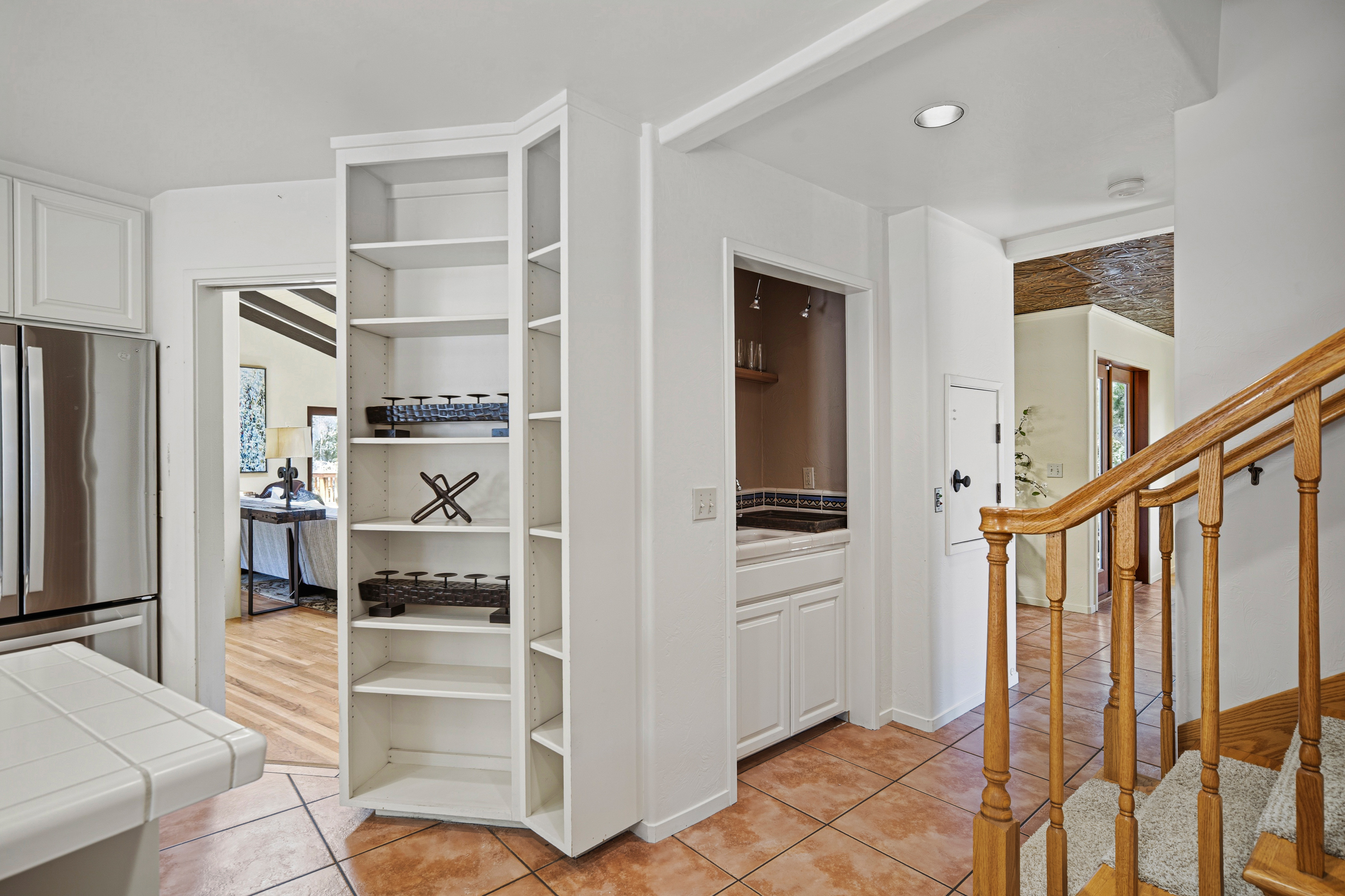 This interior view captures a transition space featuring a white corner shelving unit adjacent to a kitchen area with a stainless steel refrigerator. To the right, a small wet bar with a sink and cabinetry is tucked into a recessed alcove, leading toward a staircase with wooden railings and carpeted steps. The space is finished with warm-toned tile flooring and neutral walls, creating a functional and connected layout.