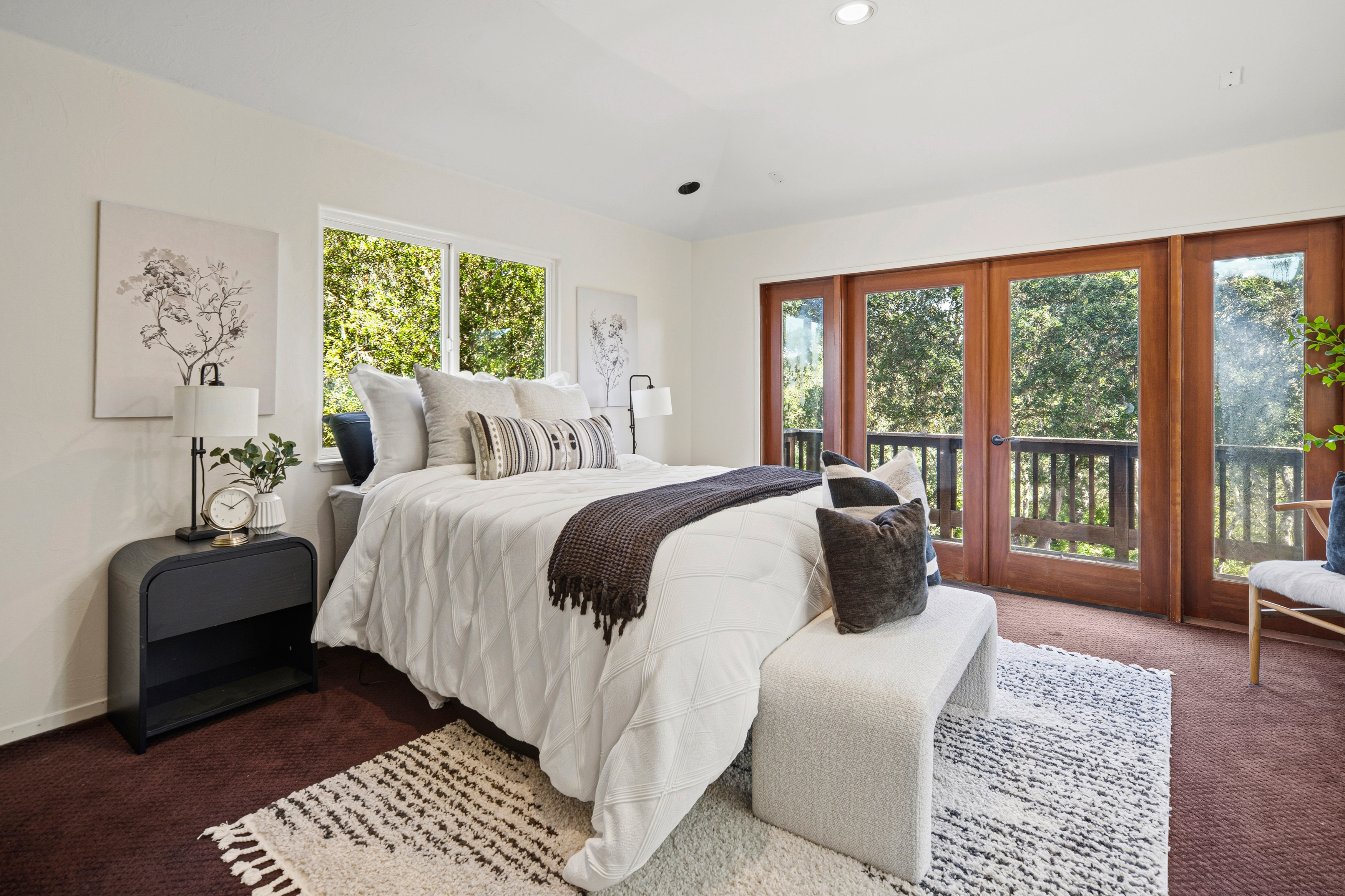 This bright and airy primary bedroom features a neutral color palette with a plush white bed dressed in textured linens and a dark throw blanket. The room is anchored by a patterned area rug and includes a modern black nightstand, a matching bench at the foot of the bed, and large glass doors that open onto a private balcony. The space is filled with natural light, creating a serene and inviting atmosphere.