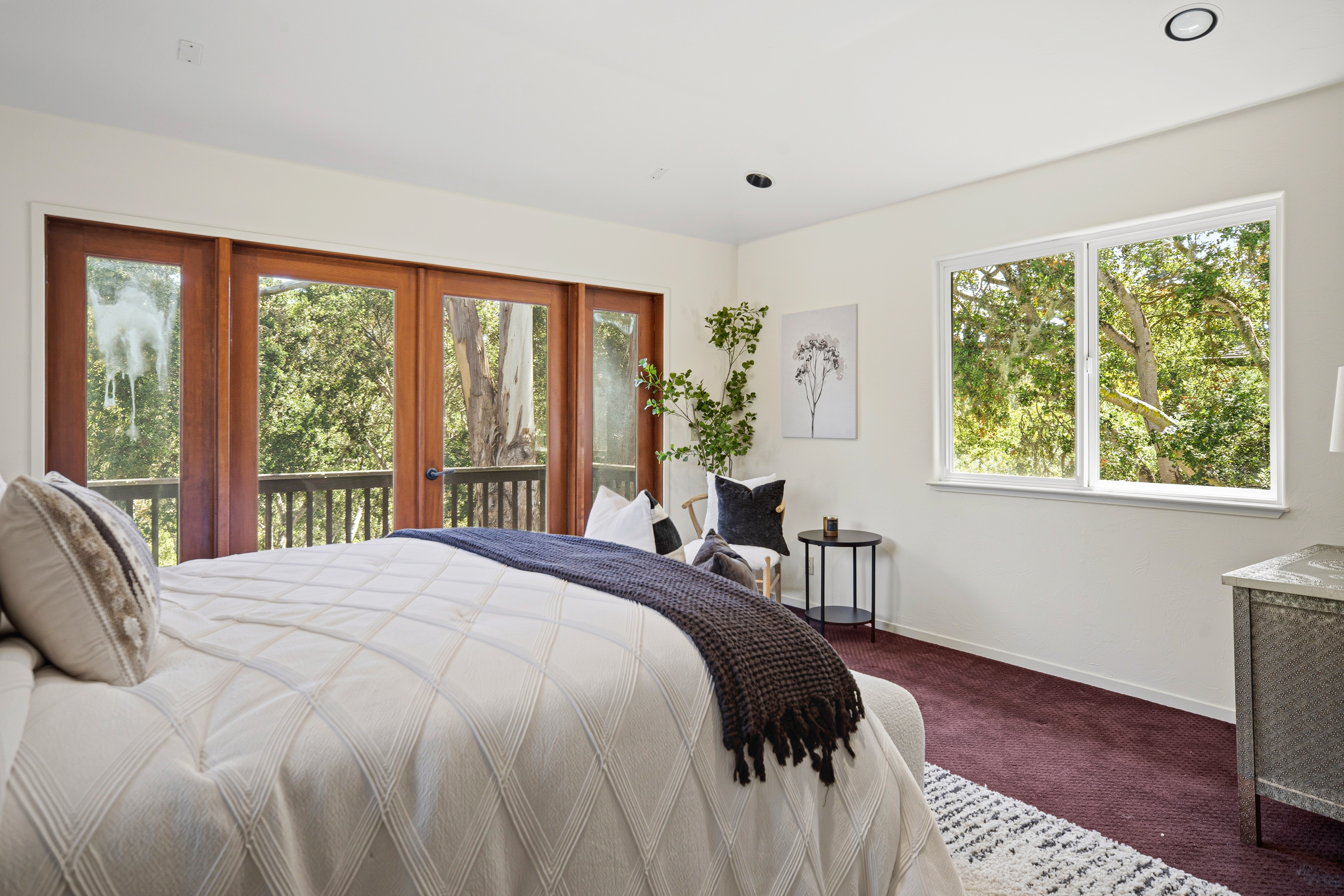 This primary bedroom features a plush bed with a textured white quilt and a dark throw blanket, positioned to face a wall of large glass doors that open onto a balcony. The room is accented by a small seating area with a round side table, a potted plant, and a piece of framed wall art. The space is finished with deep-toned carpeting and large windows that provide an expansive view of the surrounding trees.