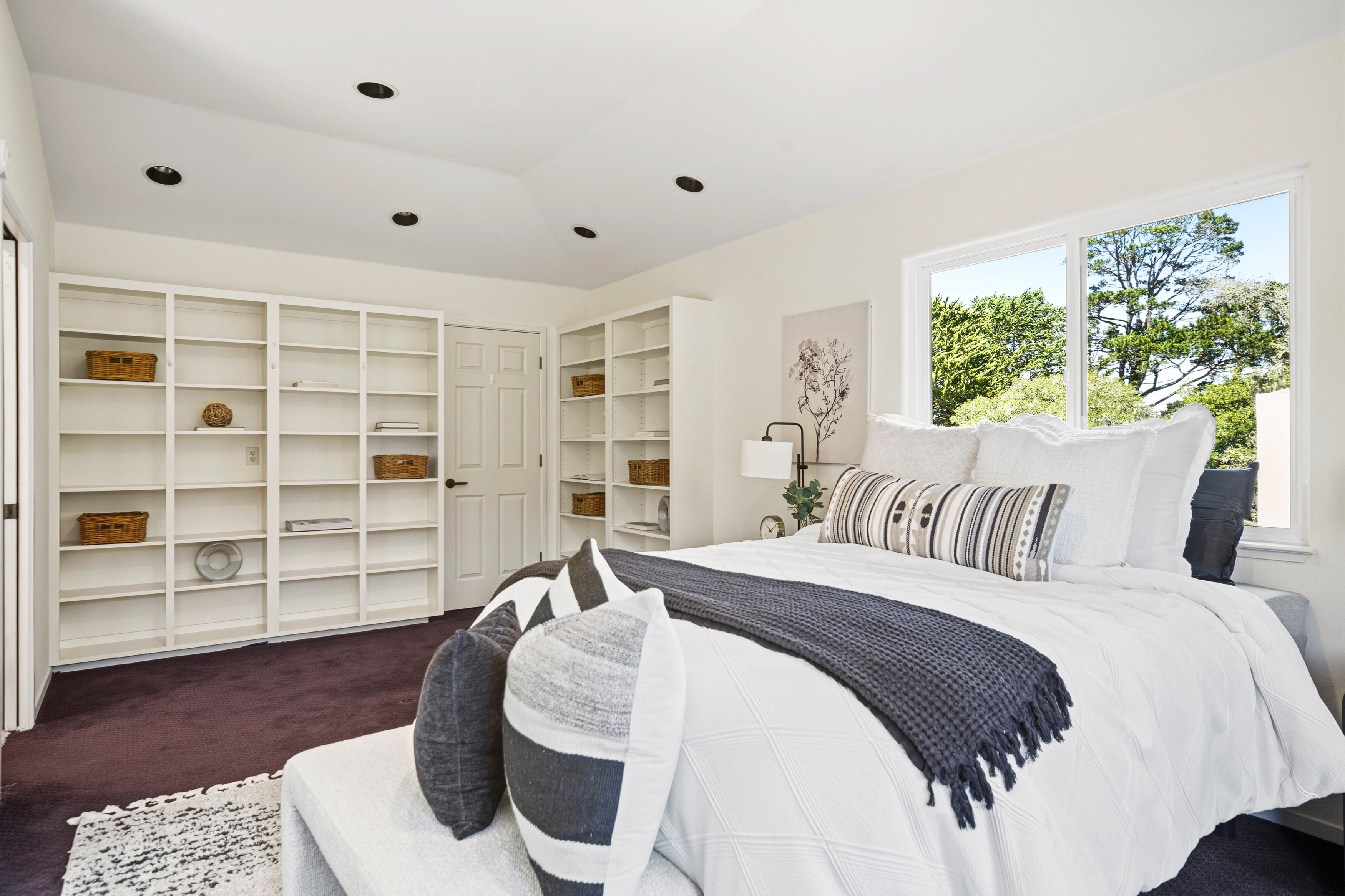 This bright and airy bedroom features a queen-sized bed dressed in crisp white linens with a dark gray throw and decorative patterned pillows. The room is accented by built-in white shelving units that provide ample storage and display space, while a large window allows for natural light and a view of the surrounding trees. The space is finished with dark carpeting and a neutral color palette, creating a calm and inviting atmosphere.