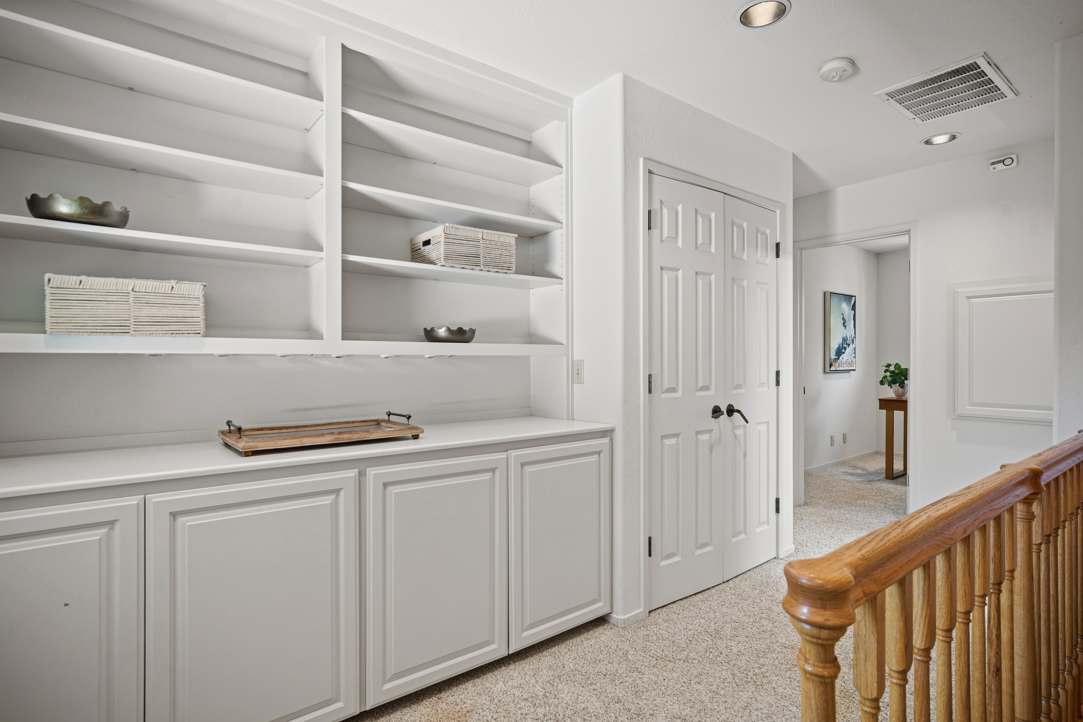 This interior view captures a bright hallway featuring built-in white cabinetry with open shelving above, providing ample storage and display space. A wooden staircase railing is visible in the foreground, leading to a carpeted corridor that opens into an adjacent room. The space is clean and neutral, with recessed lighting and a classic architectural style that creates a welcoming transition between areas.