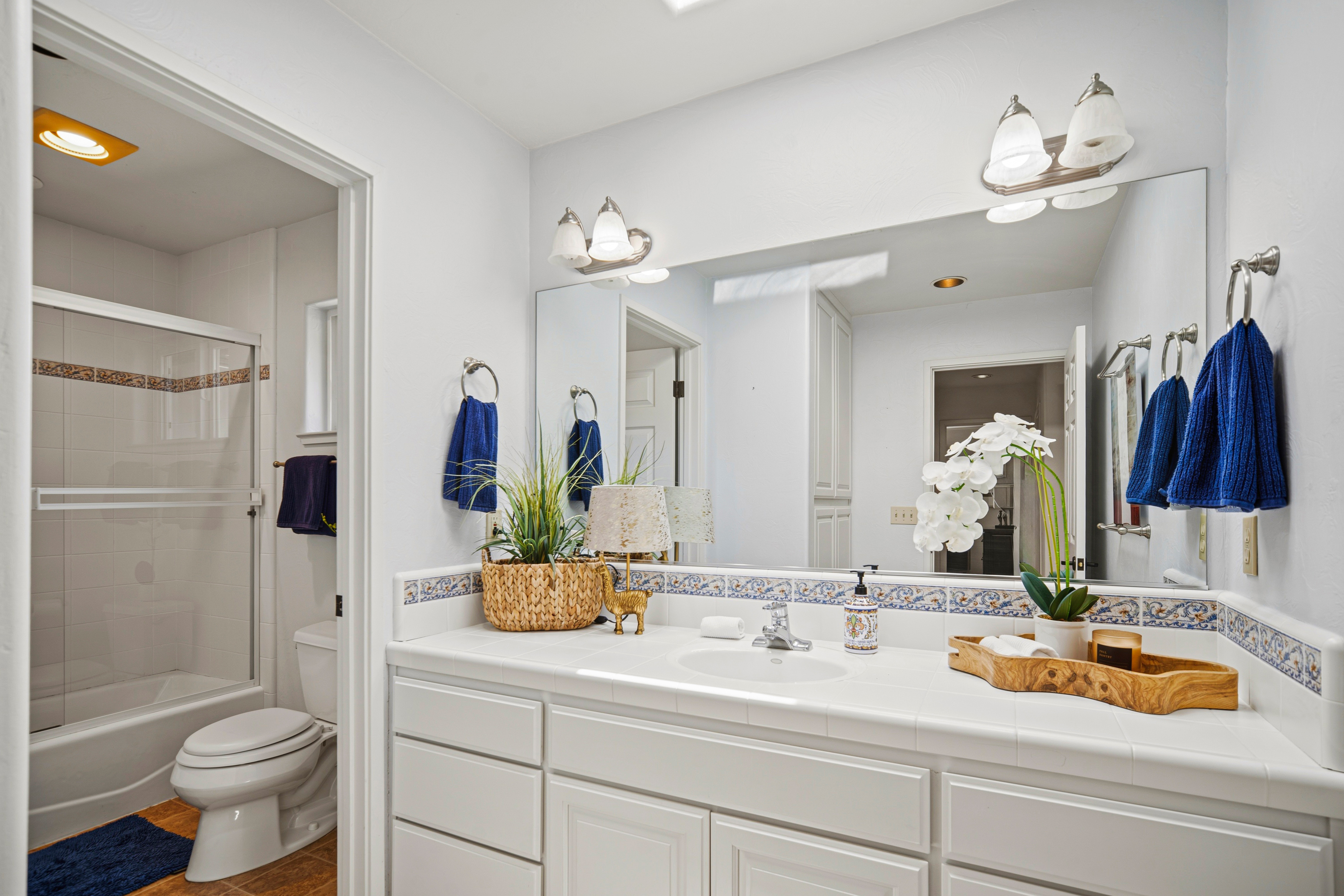 This bright primary bathroom features a white vanity with a tiled countertop and a decorative patterned tile backsplash. A large wall-mounted mirror reflects the space, which includes a separate toilet and shower area visible through an open doorway. The room is accented with blue towels and decorative greenery, creating a clean and functional aesthetic.