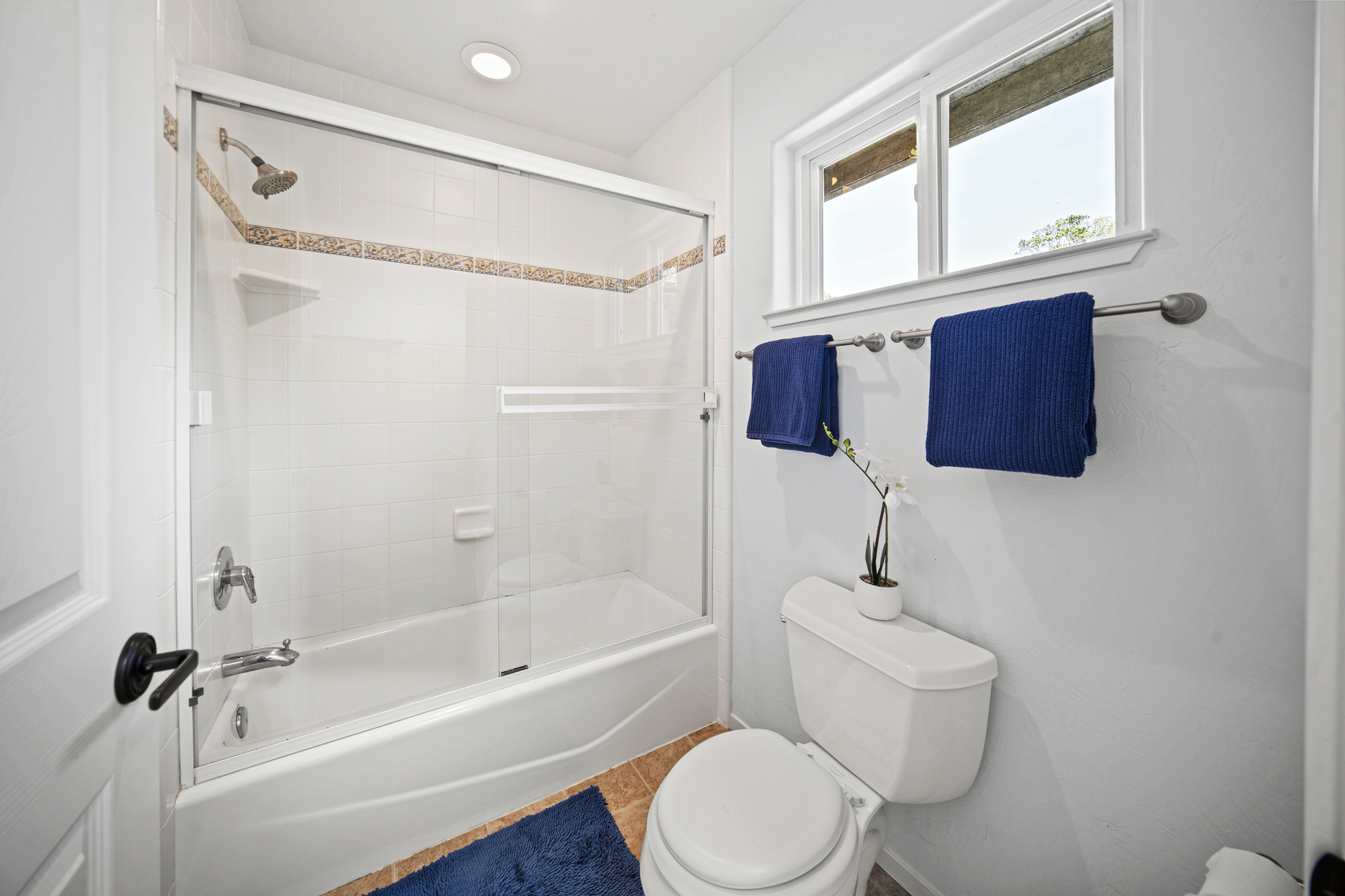 This clean and functional bathroom features a white porcelain toilet and a shower-tub combination enclosed by sliding glass doors. The walls are finished in white tile with a decorative accent border, and a window above the towel rack provides natural light. A blue rug and matching blue towels add a pop of color to the neutral space.