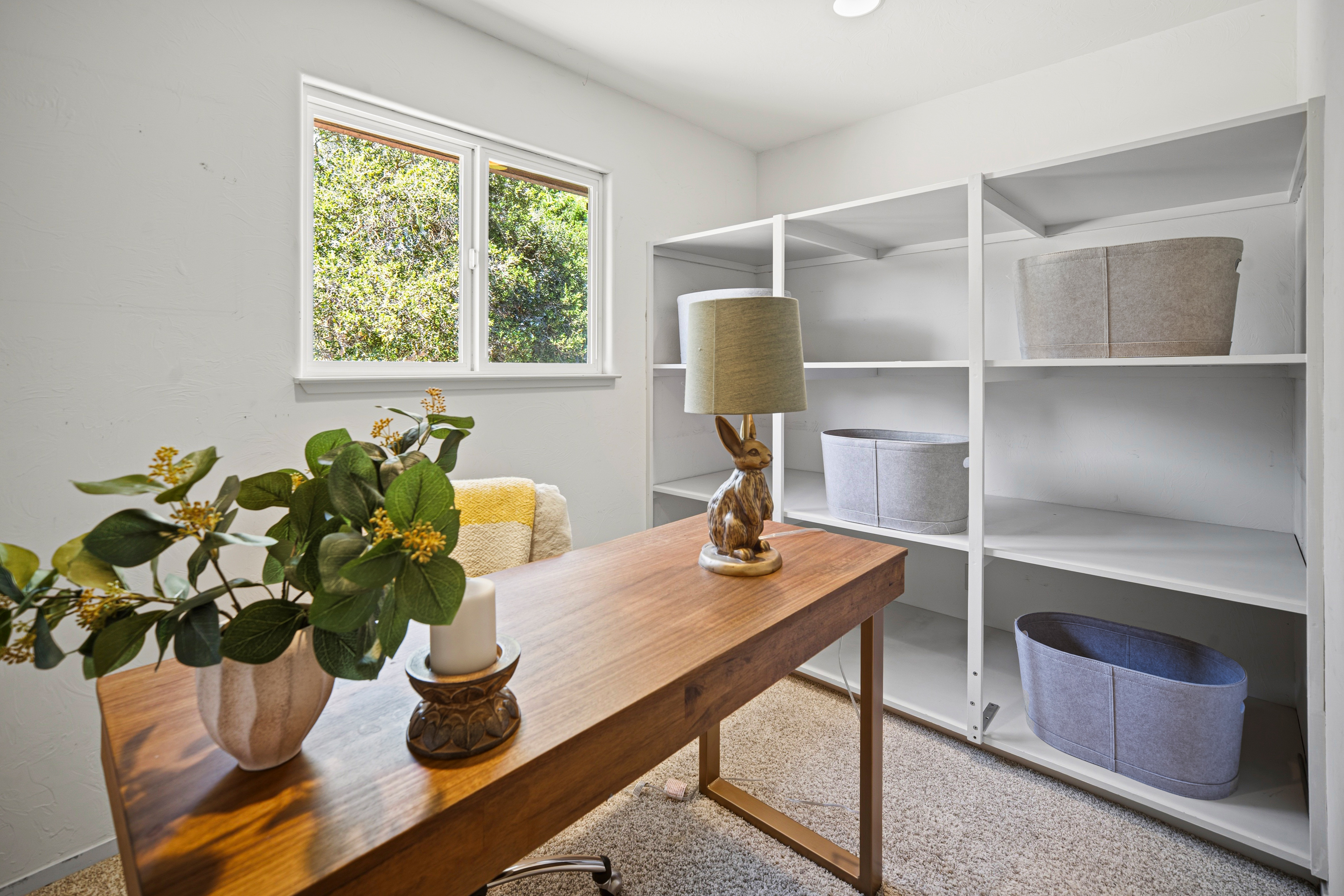 This bright home office features a warm-toned wooden desk centered in the room, accented by a whimsical rabbit-shaped table lamp and a potted plant. A large white shelving unit with grey felt storage bins provides ample organizational space against the wall. The room is illuminated by natural light from a window overlooking greenery, creating a clean and functional workspace.