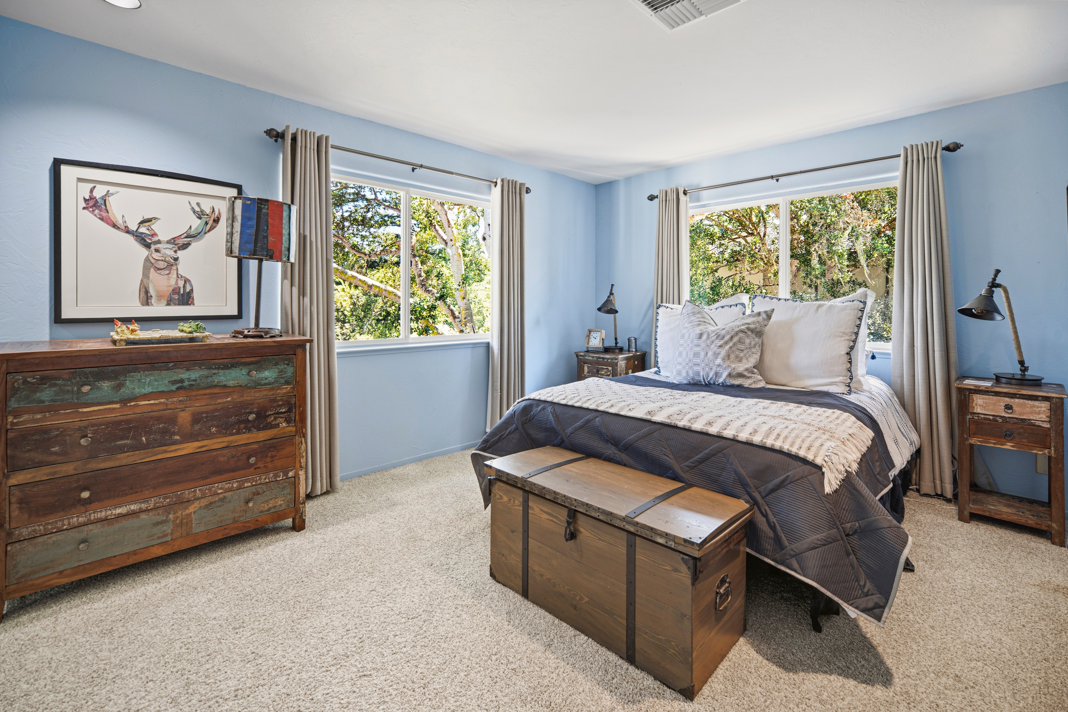This bedroom features light blue walls, neutral carpeting, and a rustic aesthetic highlighted by a distressed wood dresser and a matching wooden storage chest at the foot of the bed. Two windows with light-colored curtains provide natural light and views of the surrounding trees, while a framed deer print adds a touch of character to the space. The room is furnished with a bed dressed in a dark blue quilt and a pair of bedside tables with matching lamps, creating a cozy and inviting atmosphere.