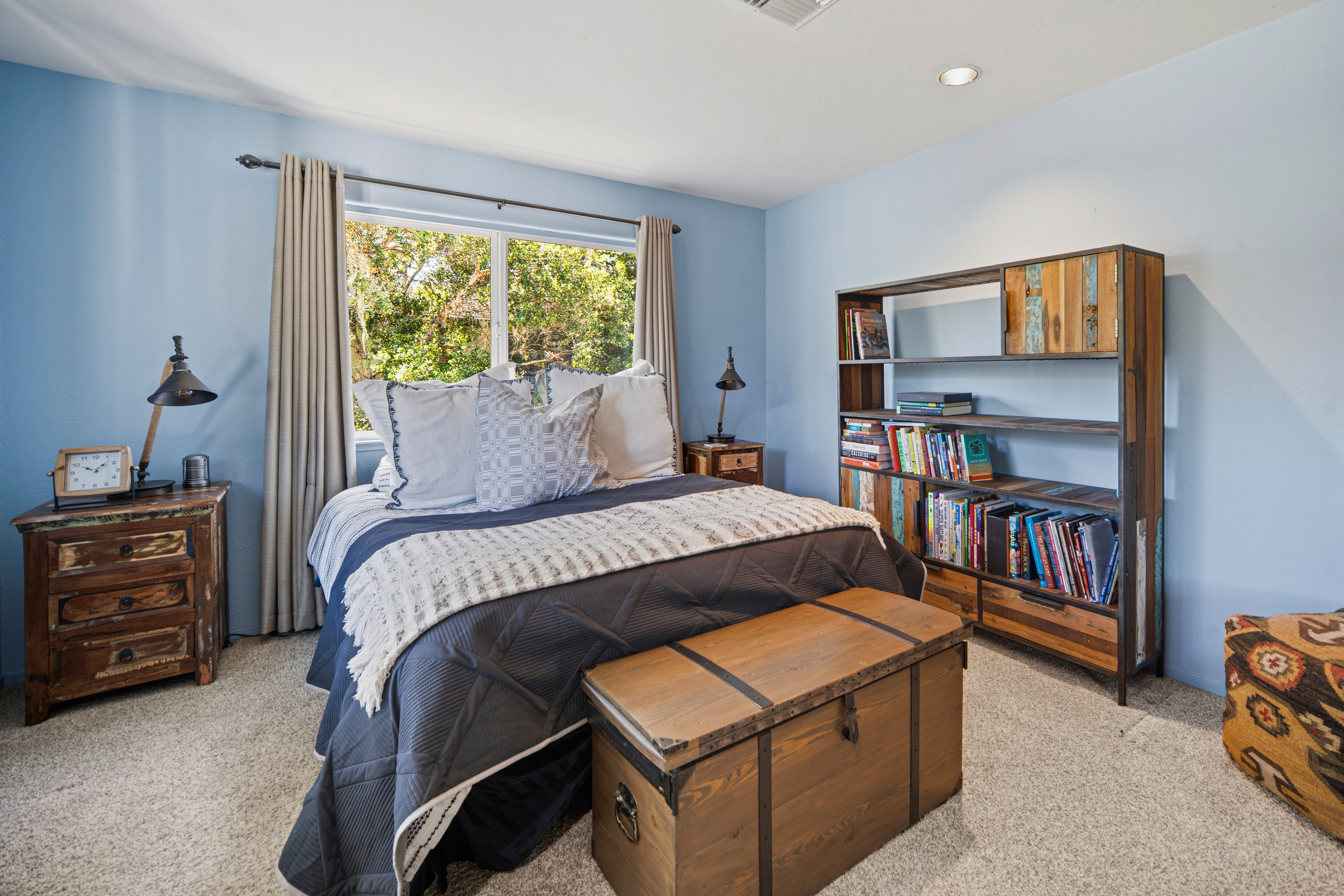 This cozy bedroom features light blue walls and a rustic aesthetic, centered around a bed dressed in a dark quilted comforter and a patterned throw. The room is furnished with matching dark wood nightstands, a large wooden storage chest at the foot of the bed, and a tall, multi-shelf bookcase filled with books. A large window with neutral curtains allows natural light to brighten the space, which is finished with light-colored carpeting.