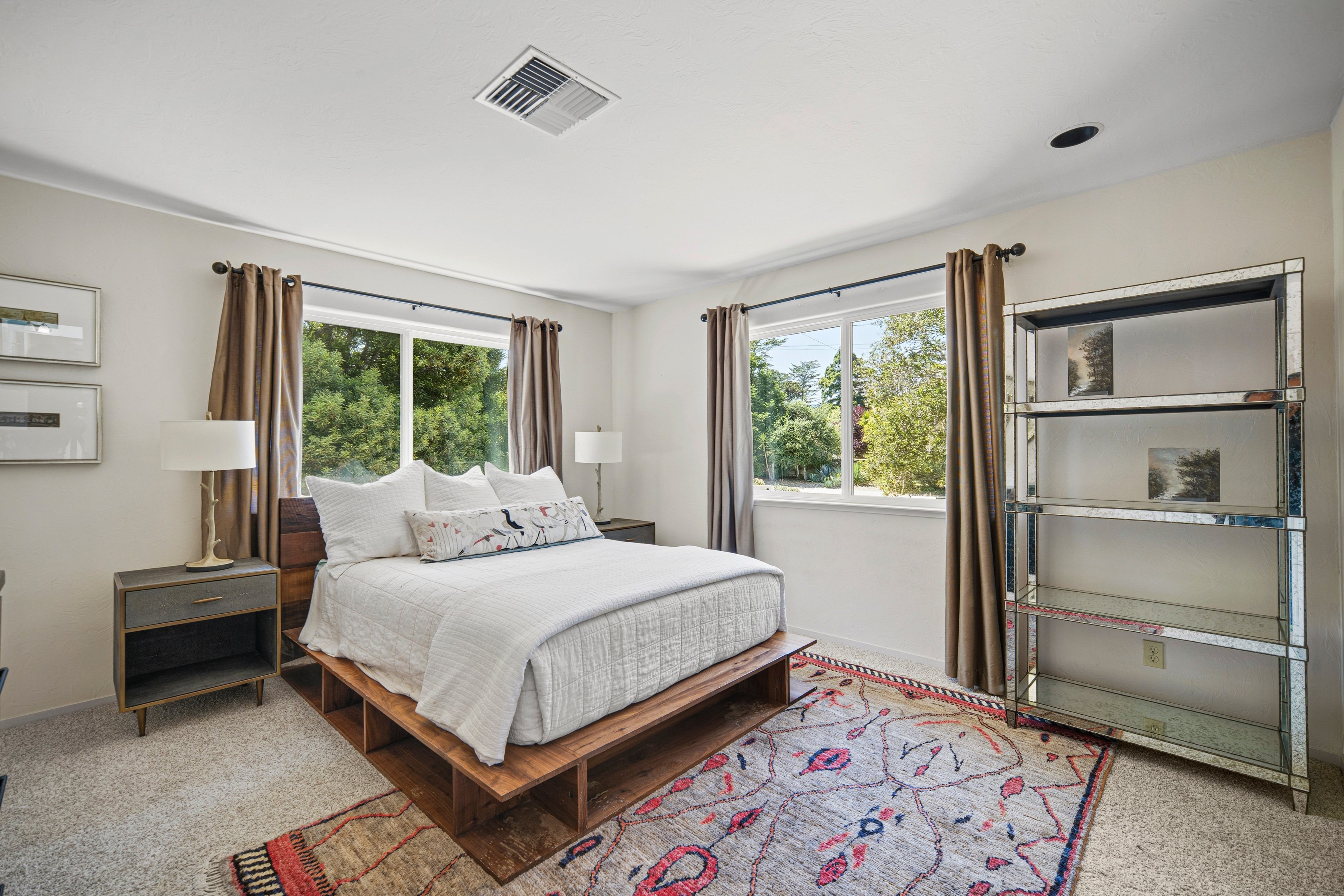 This bright and airy bedroom features a low-profile wooden platform bed frame centered between two windows dressed in brown curtains. To the right, a tall, mirrored shelving unit adds a touch of modern elegance, while a patterned area rug anchors the space over neutral carpeting. The room is completed by two bedside tables with matching lamps and framed artwork on the wall, creating a clean and inviting atmosphere.