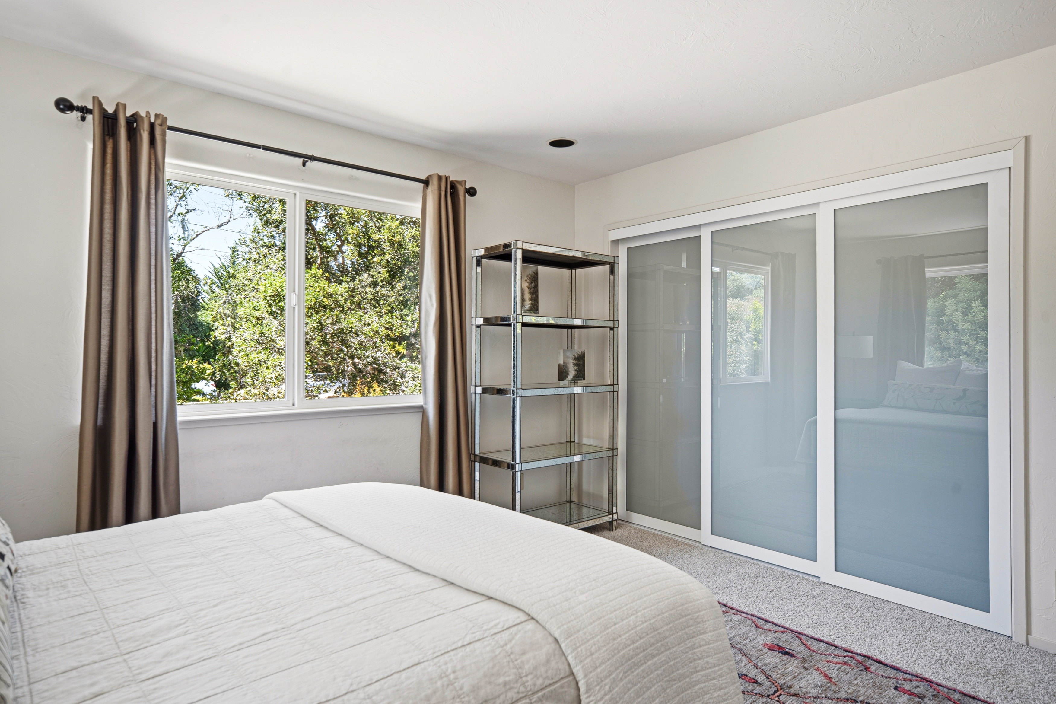 This bright bedroom features a neutral color palette with a white bedspread and soft grey carpeting. A large window with brown curtains allows natural light to fill the room, while a tall, metallic shelving unit stands beside a set of sliding glass closet doors. The space offers a clean, minimalist aesthetic with a view of lush greenery outside.
