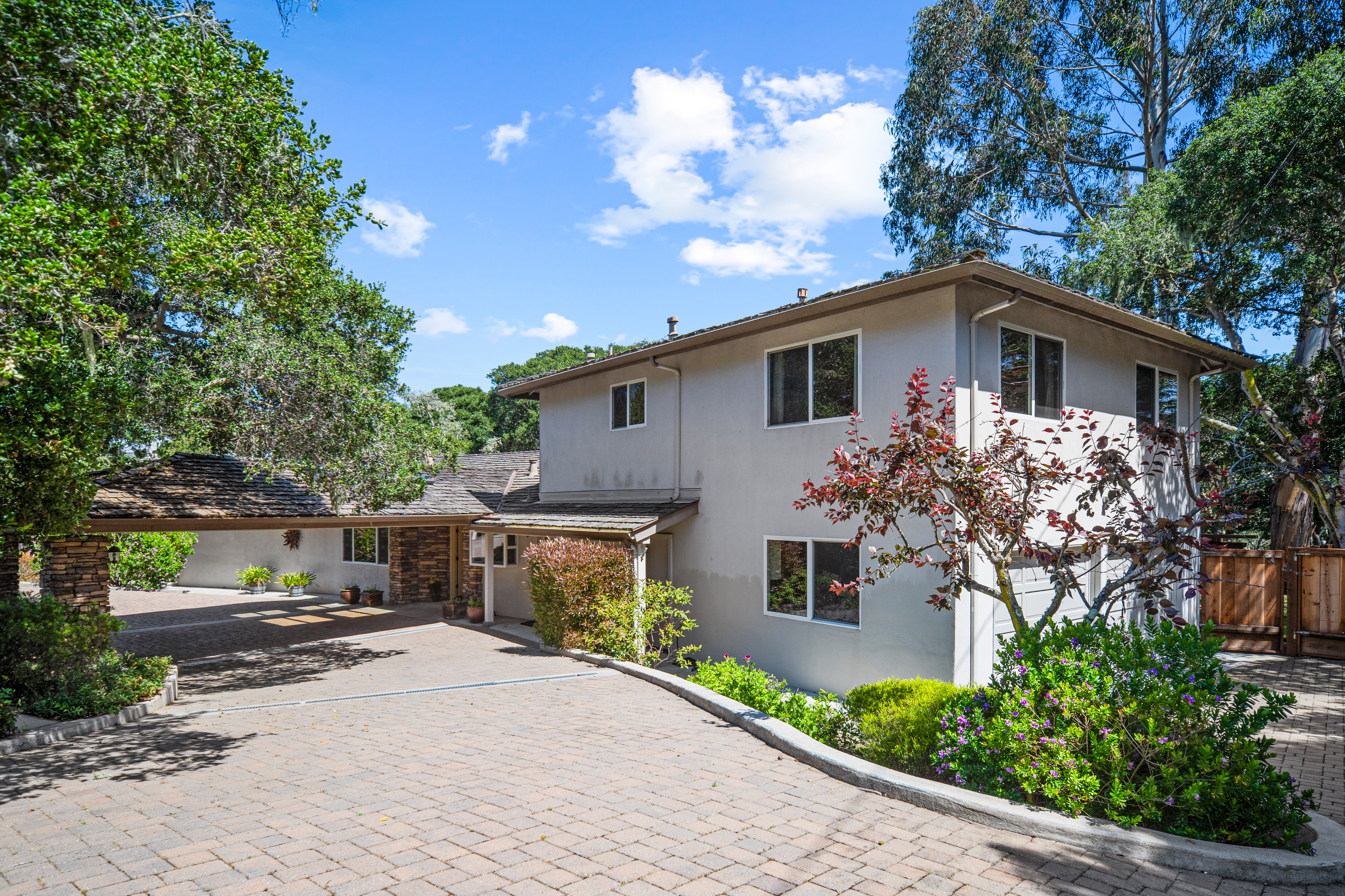 This two-story home features a light-colored stucco exterior and a sprawling brick-paved driveway leading to a covered carport. The property is surrounded by mature trees and lush landscaping, creating a private and serene atmosphere. The perspective captures the home's inviting curb appeal, highlighting the architectural blend of the main structure and the extended carport area.