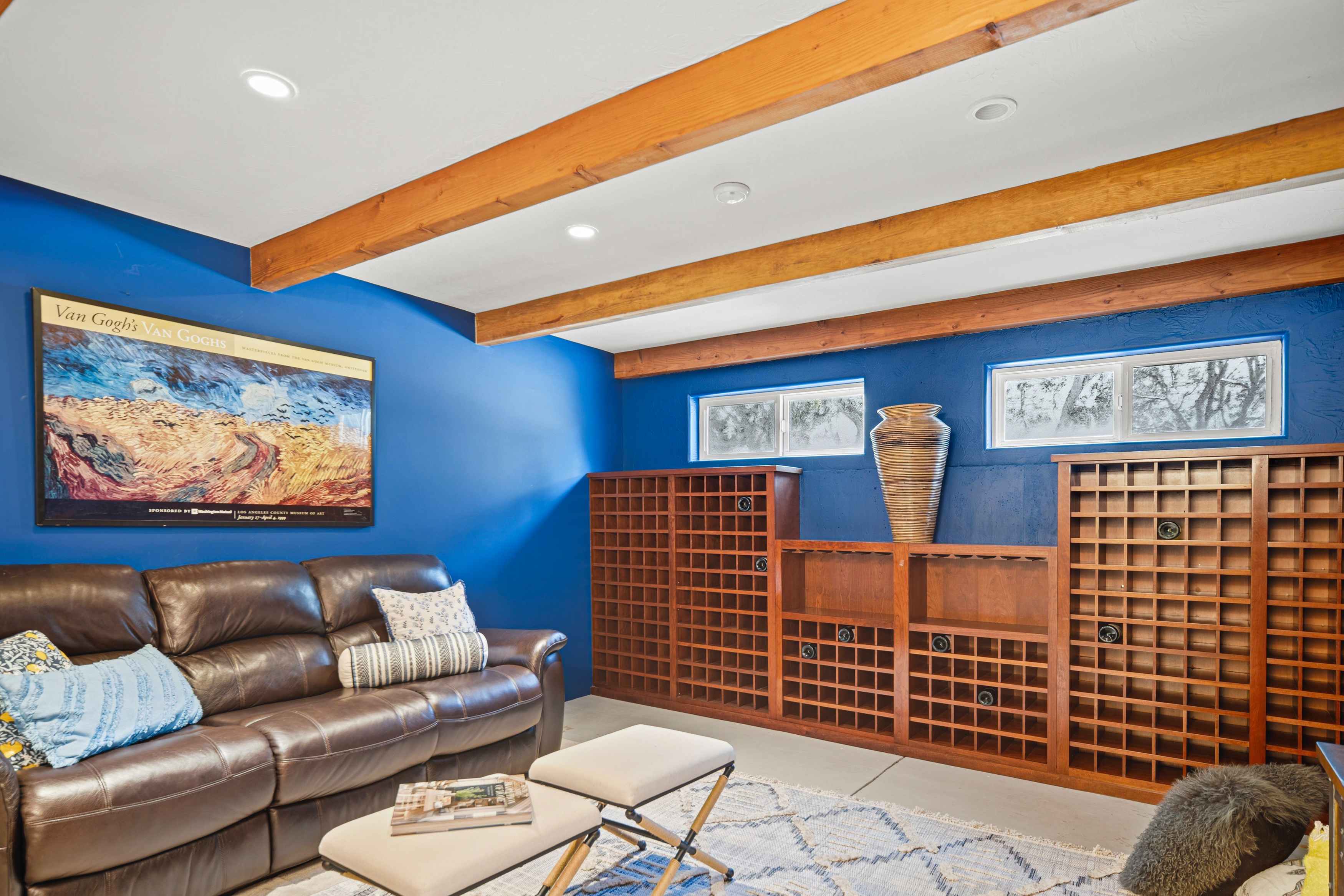 This cozy basement living area features vibrant blue walls, exposed wood ceiling beams, and a large brown leather sofa. A custom wooden wine storage unit spans the back wall, complemented by a decorative vase and two small windows that provide natural light. The space is grounded by a patterned area rug and two ottomans, creating a comfortable and inviting atmosphere.