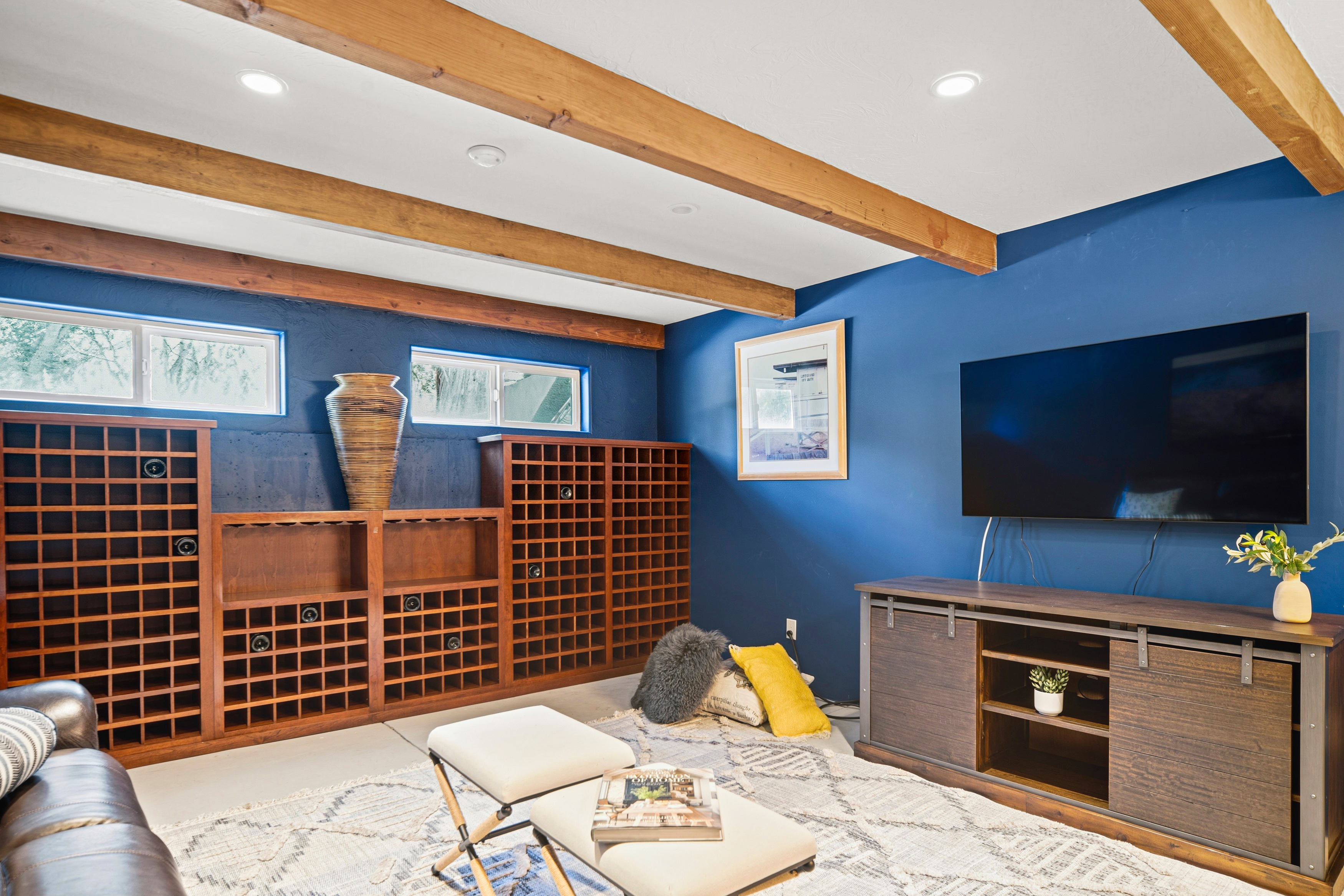This cozy basement living area features a bold blue accent wall, exposed wooden ceiling beams, and a large wall-mounted television. A custom wooden wine rack system spans the back wall, complemented by a sliding barn-door style media console and a patterned area rug. The space is furnished with a leather sofa and two ottomans, creating a comfortable and inviting atmosphere for relaxation or entertainment.