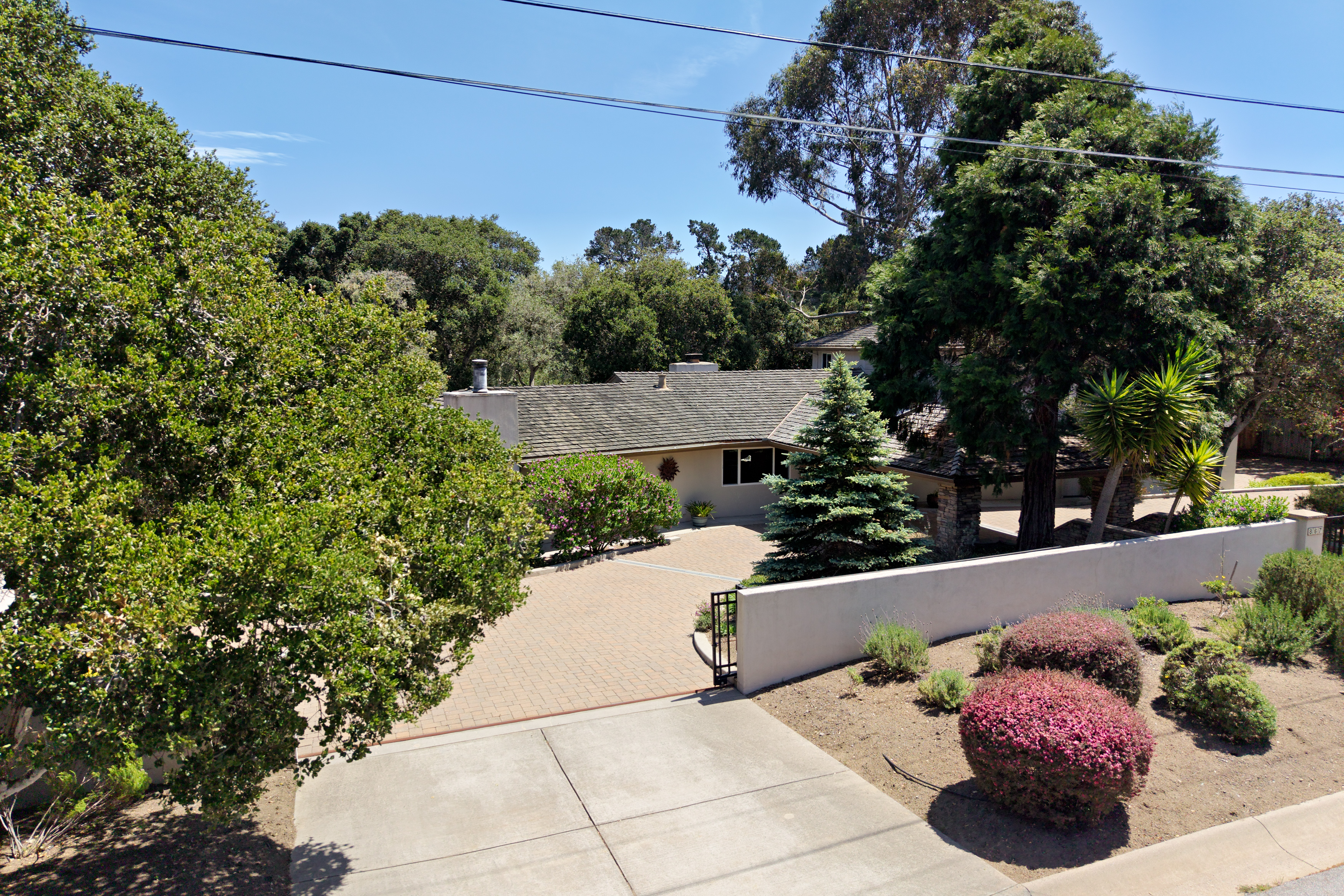 This elevated perspective captures the front exterior of a single-story residence, showcasing a paved driveway leading to a gated entrance. The home features a neutral-toned facade, a shingled roof, and mature landscaping, including a prominent evergreen tree and vibrant flowering shrubs. The scene conveys a sense of privacy and established curb appeal, framed by lush trees under a clear blue sky.