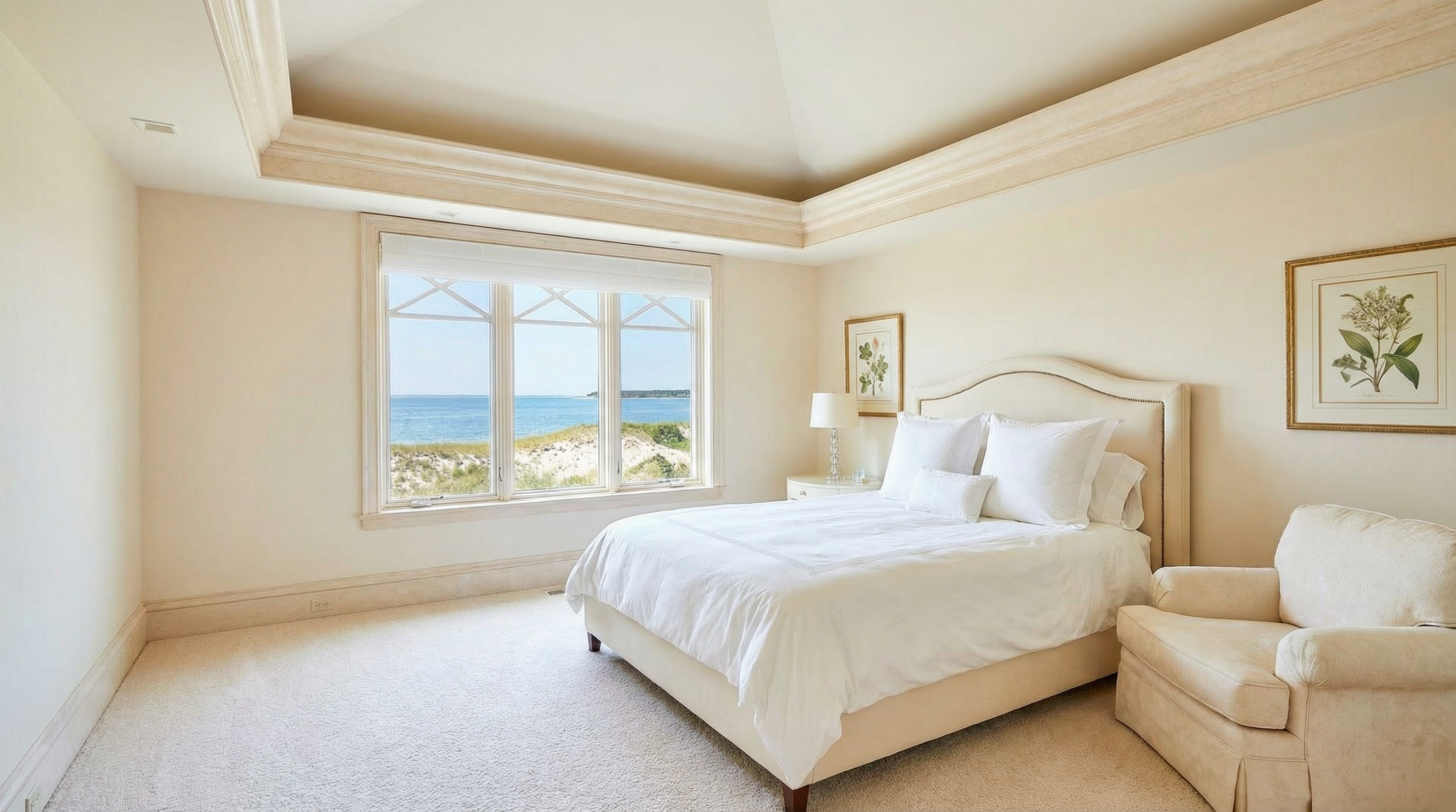 This is a bright and airy primary bedroom, featuring a large window with a view of the ocean. The room is decorated in neutral tones, with white bedding and a beige armchair. The architectural details include a tray ceiling with molding, which adds a touch of elegance.