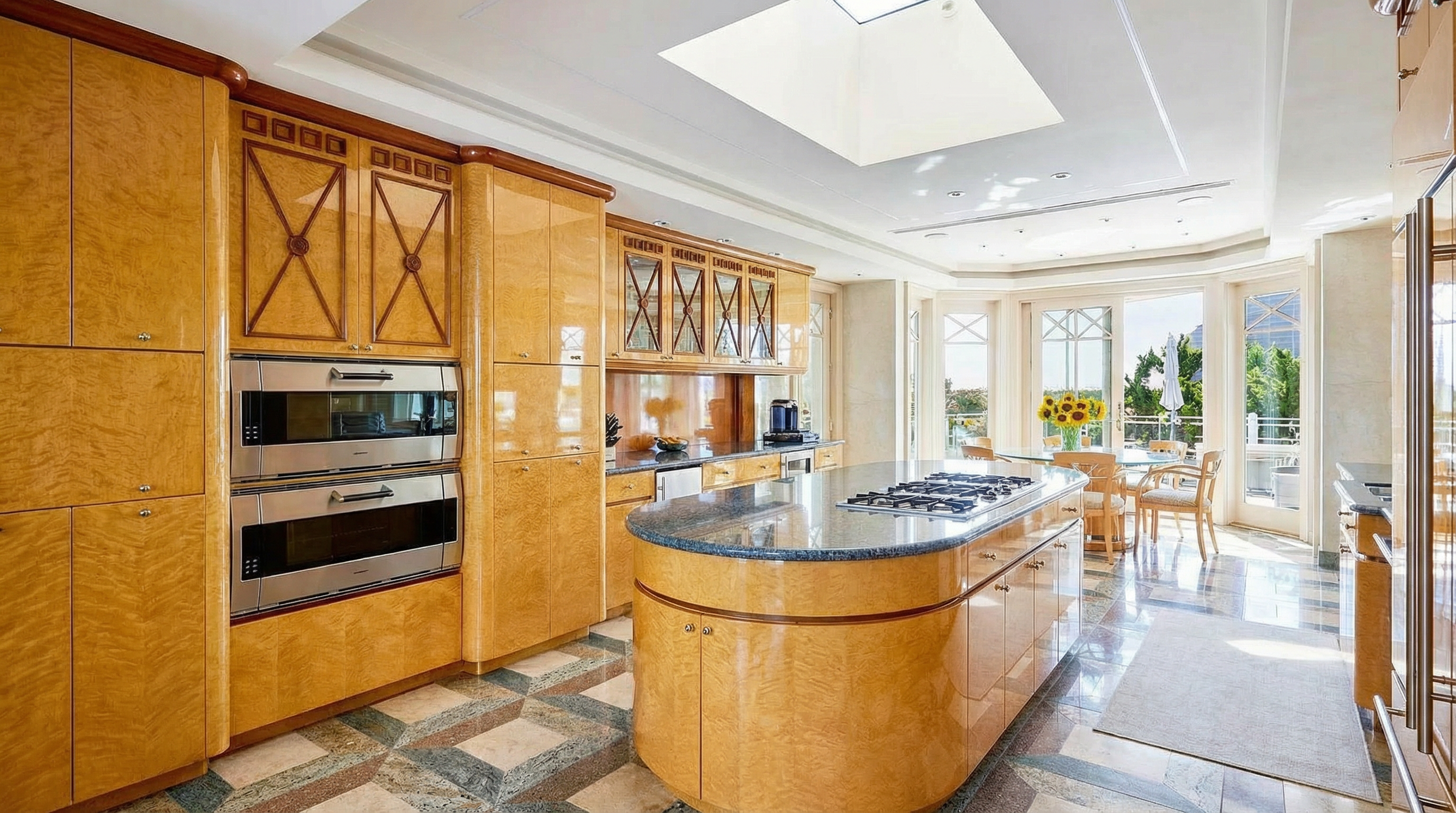 This is a well-appointed kitchen with custom wood cabinetry and high-end appliances. The space features a large, curved island with a cooktop, providing ample workspace. A light-filled breakfast nook extends the kitchen area, offering a pleasant dining space with views to the exterior.