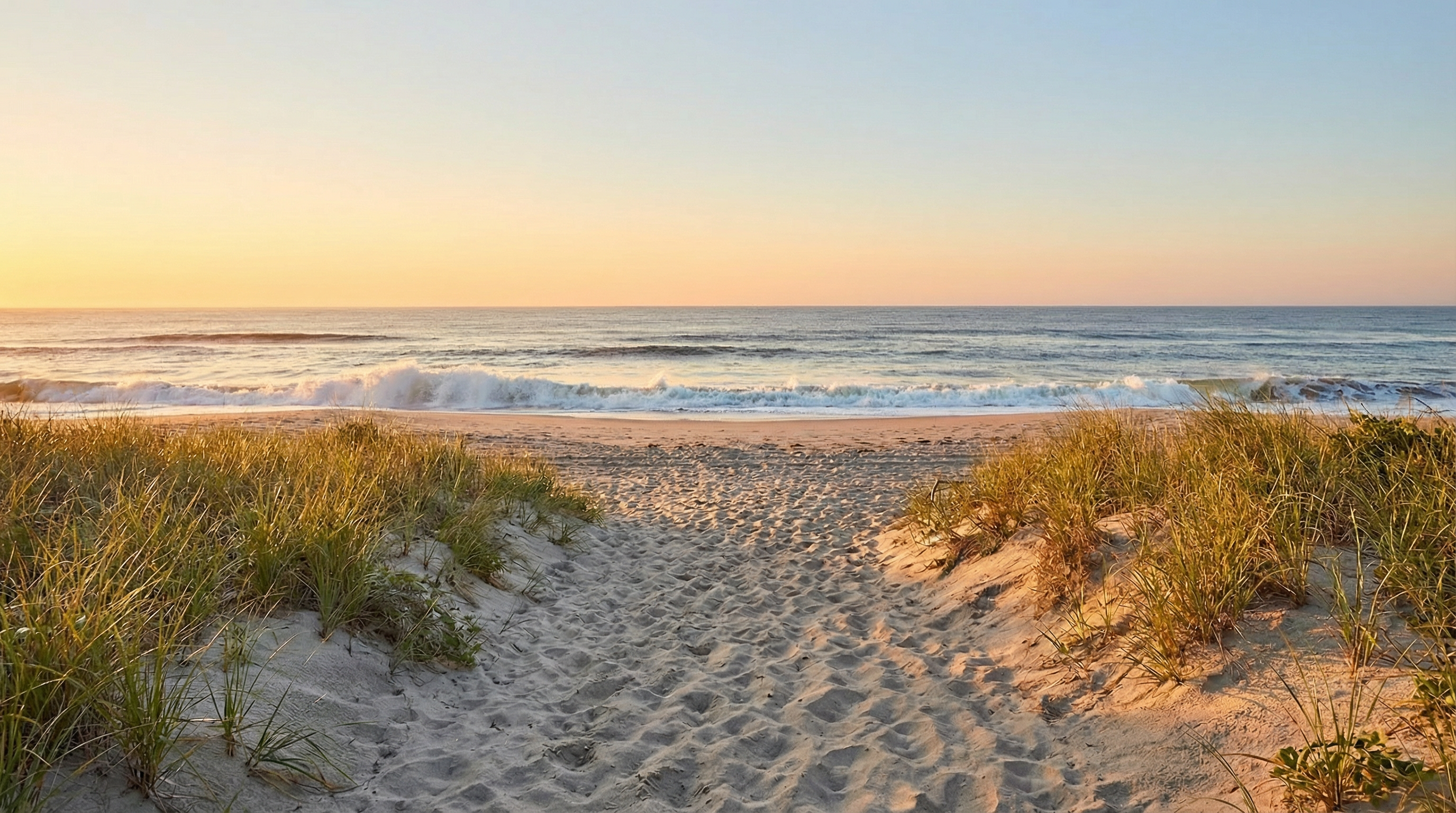 This picturesque yard features a sandy path leading from the beach to the water. Beach grass flanks the path, creating a natural border and adding to the serene atmosphere. The gentle waves and clear sky suggest a tranquil and desirable coastal setting, appealing to buyers seeking a relaxing outdoor space.