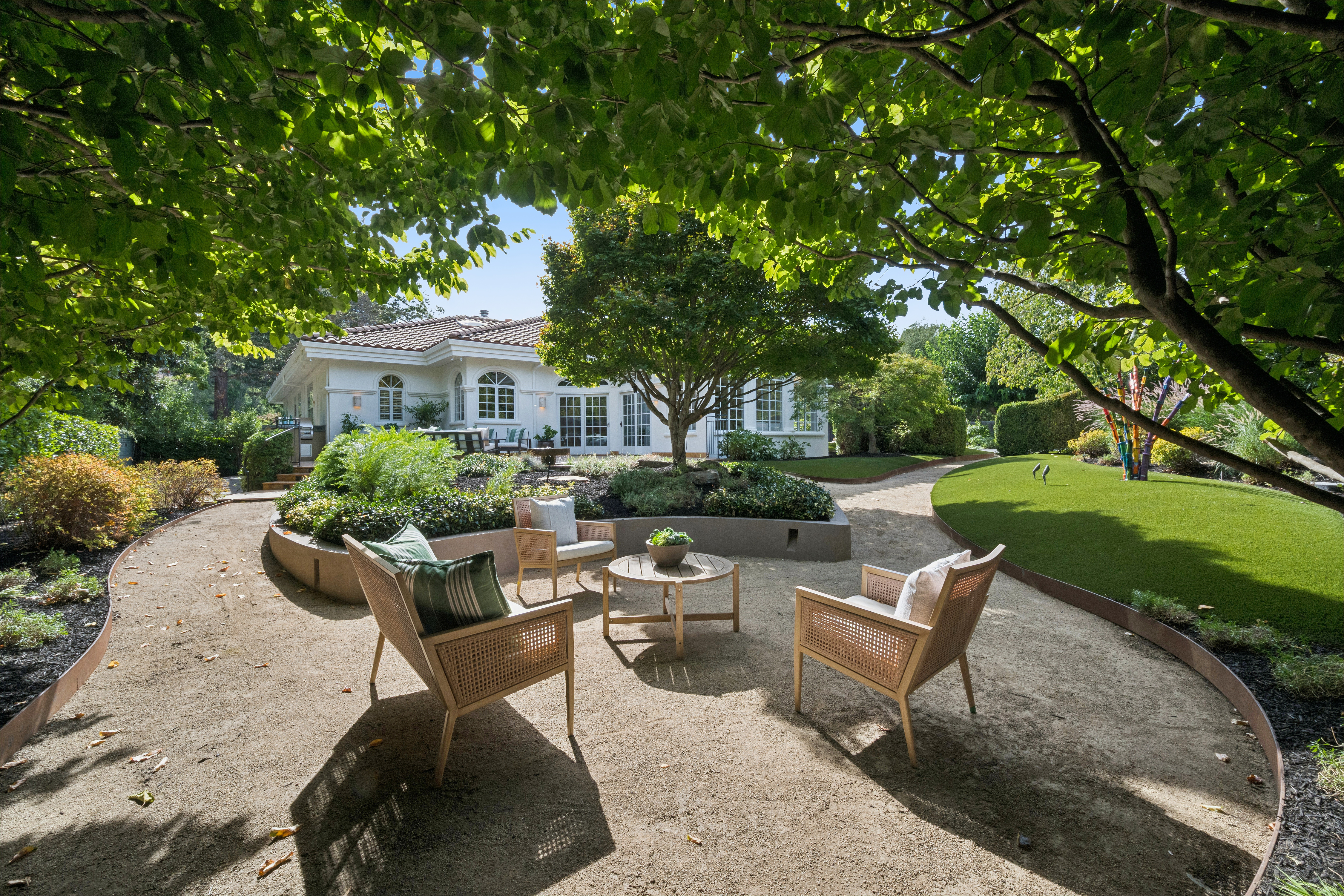 This serene backyard features a beautifully landscaped garden with a central seating area nestled in a gravel courtyard, perfect for outdoor relaxation. Lush greenery and mature trees provide dappled shade over the space, complemented by a neatly manicured lawn and a sophisticated raised garden bed. The scene is anchored by a charming, light-colored home with classic architectural details, creating an inviting and peaceful retreat.