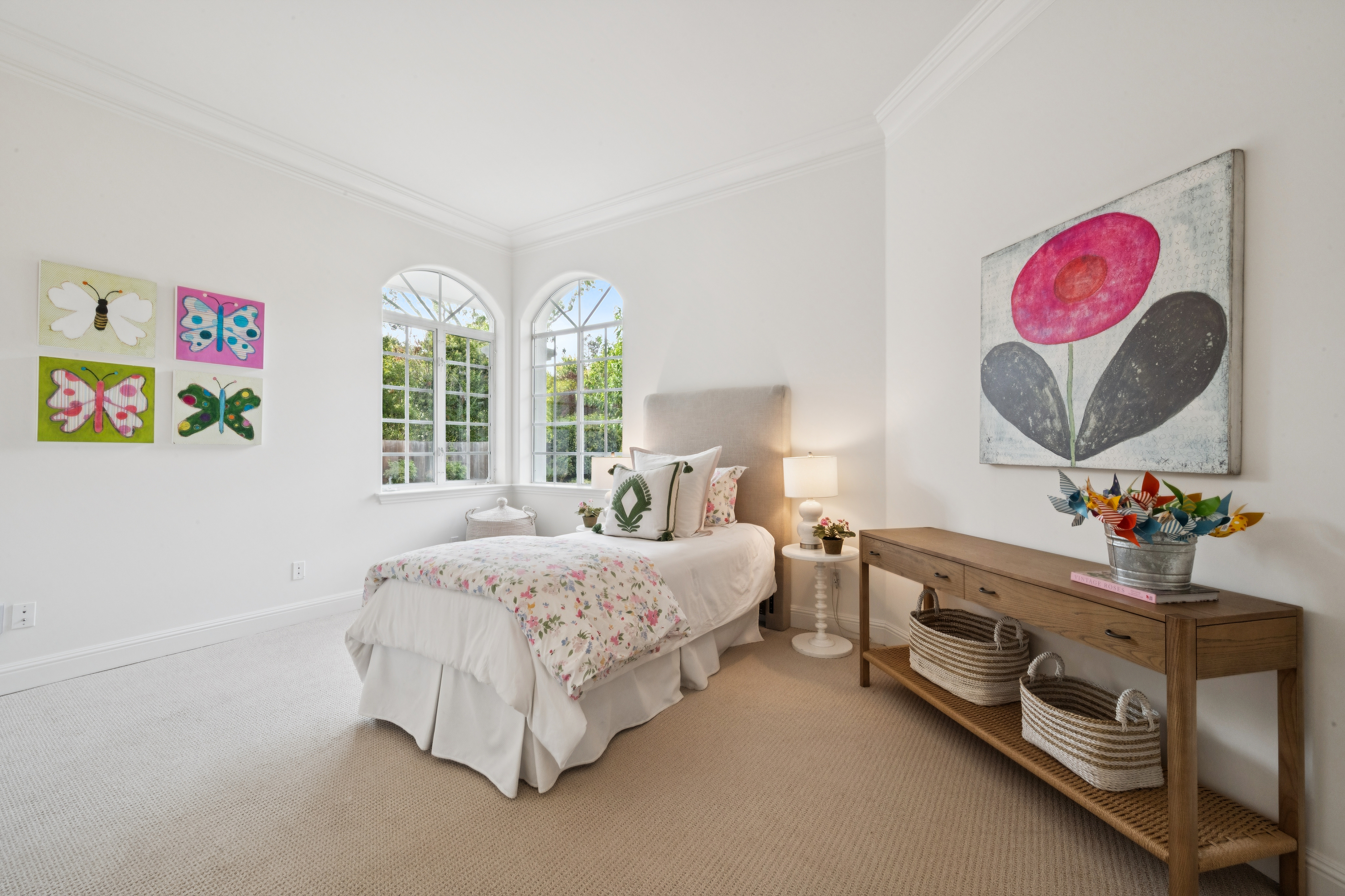 This charming and well-lit bedroom features soft, neutral carpeting and pristine white walls, accented by colorful butterfly art and a playful floral-print duvet. A tall, upholstered headboard anchors the space, while a wooden console table with woven baskets offers convenient storage. Two arched windows flood the room with natural light, creating a bright and inviting atmosphere perfect for a guest space or child's room.