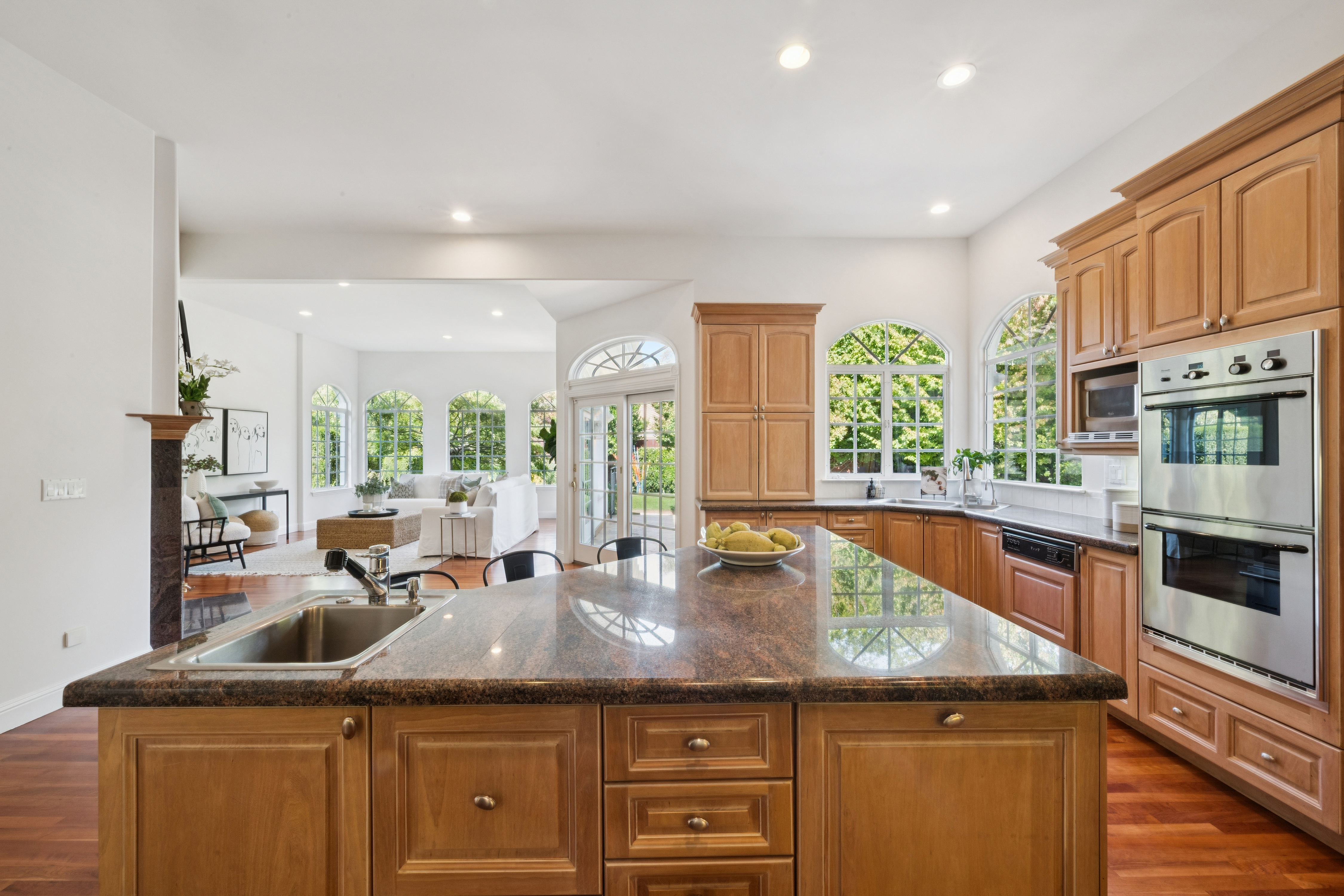 This spacious kitchen features warm wood cabinetry, granite countertops, and a large central island that anchors the room. The layout flows seamlessly into a bright living area, creating an open and inviting atmosphere with ample natural light from arched windows. The combination of hardwood flooring and professional-grade stainless steel appliances offers both elegance and functional appeal.