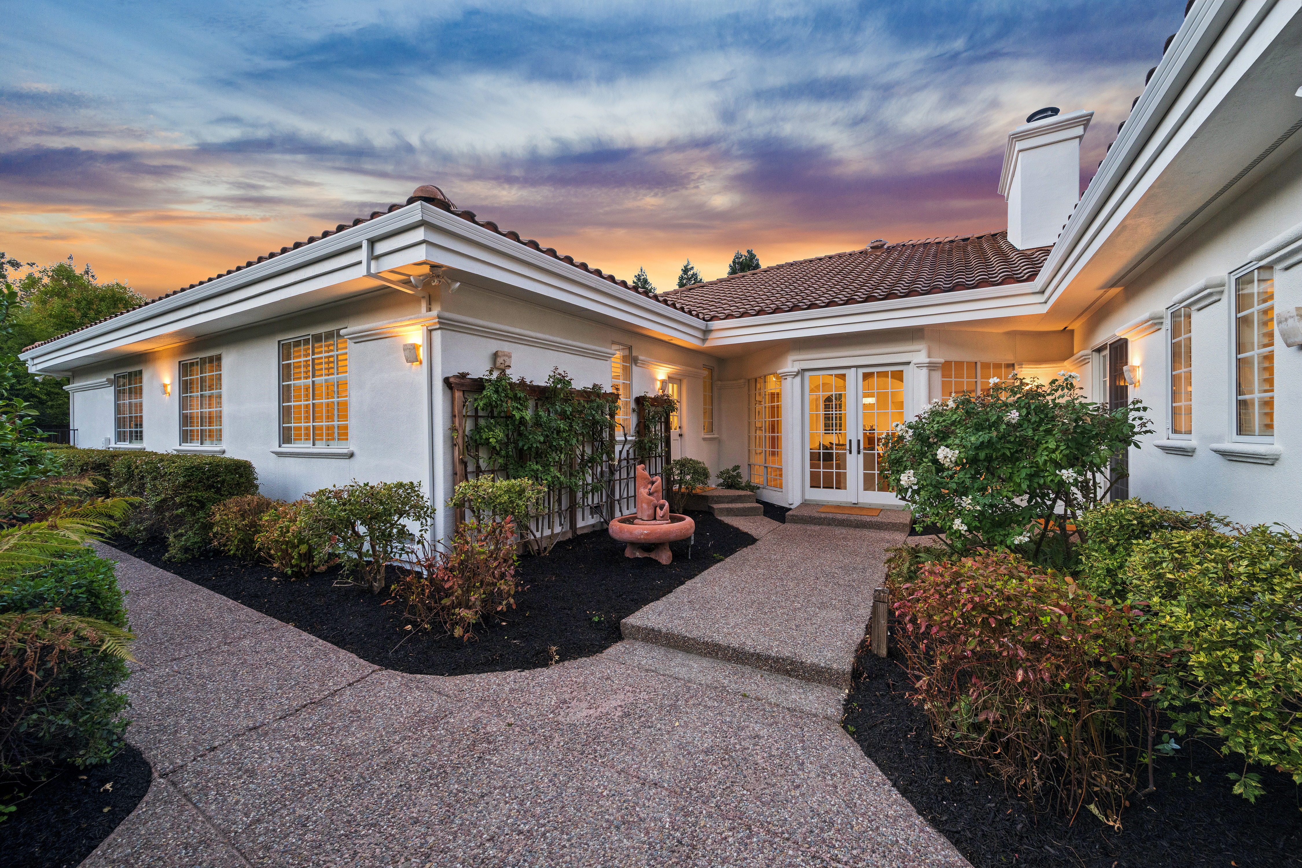 This rear view of a single-story residence showcases a well-maintained exterior with classic terracotta roof tiles, clean white stucco walls, and large windows that provide a warm, inviting glow from within. A textured concrete walkway leads to a set of French doors, framed by manicured landscaping, mature shrubs, and a small decorative fountain in a dark mulch bed. The evening sky with its vibrant sunset colors enhances the peaceful, charming aesthetic of this backyard patio area.