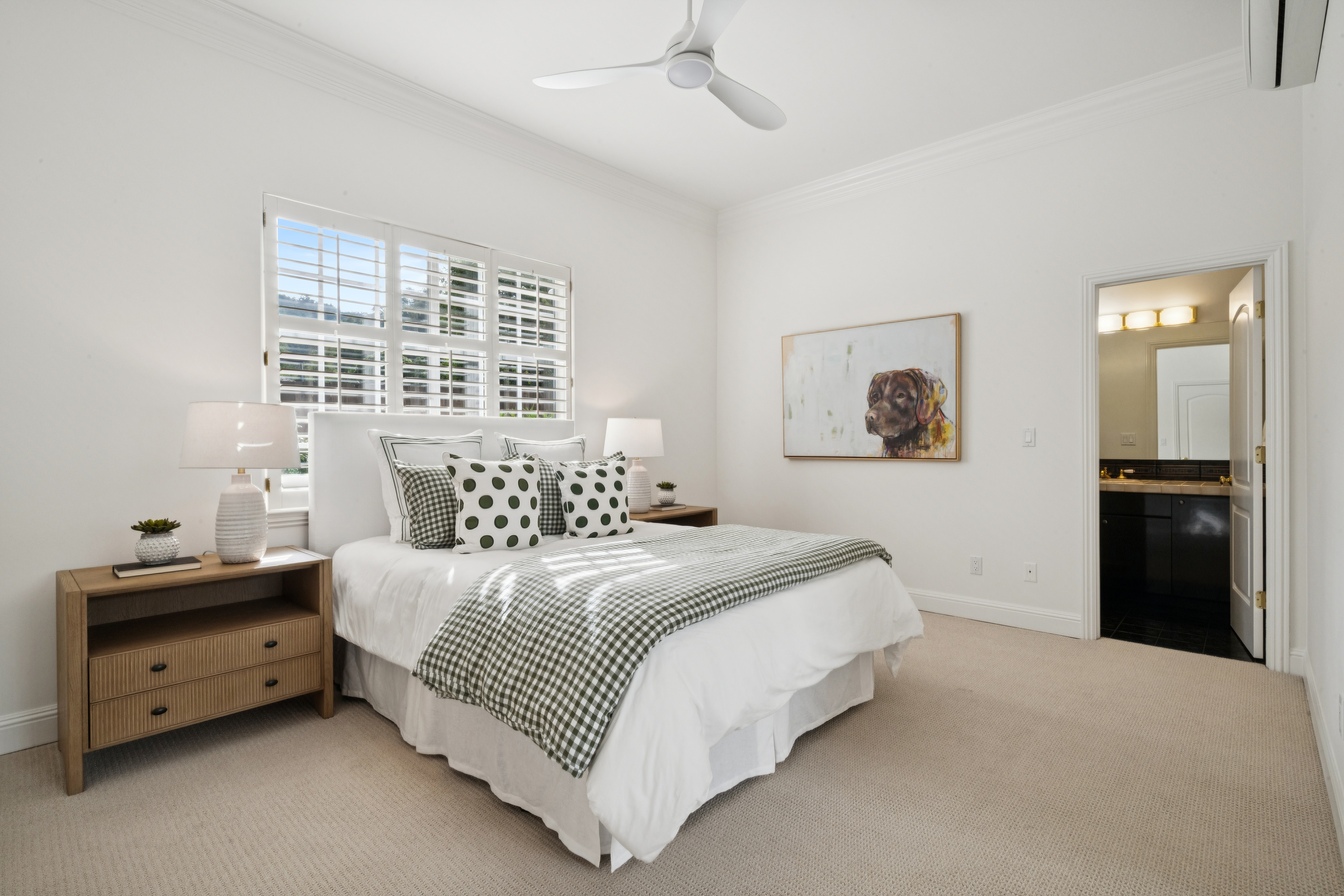 This bright and inviting bedroom features a queen-sized bed adorned with green-and-white checkered bedding and playful polka-dot accent pillows. The space is complemented by natural wood nightstands, white plantation shutters on the window, and a whimsical dog painting, creating a clean, cozy, and well-designed retreat. A doorway leads directly into an attached bathroom, adding convenience and functionality to this tranquil primary suite.