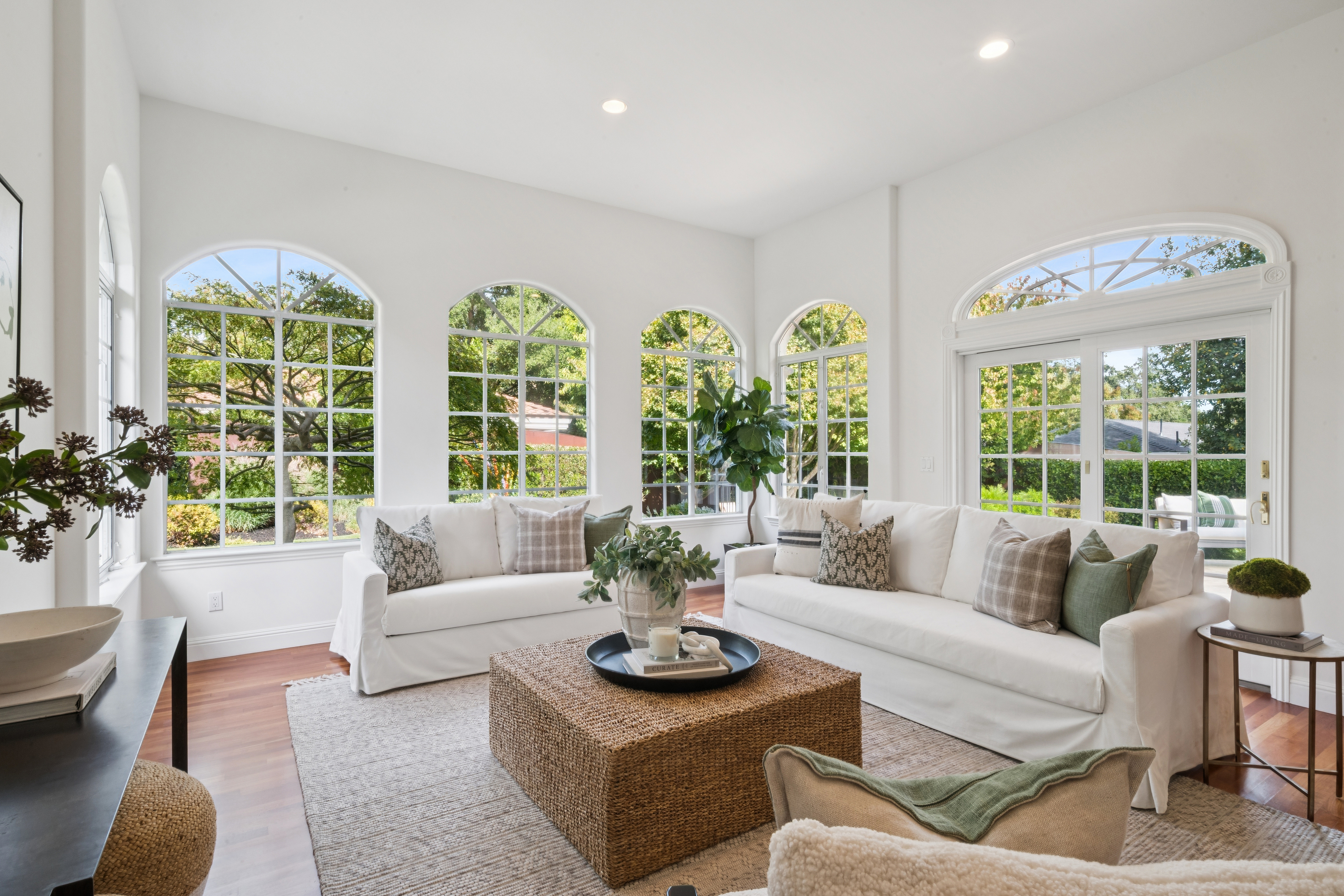 This bright and inviting living room features a spacious, neutral-toned seating arrangement anchored by a large woven square coffee table. Natural light floods the space through multiple large arched windows and a set of French doors, creating an airy and relaxed atmosphere. Rich hardwood flooring and carefully curated decorative accents contribute to a clean, transitional aesthetic suitable for comfortable family living.