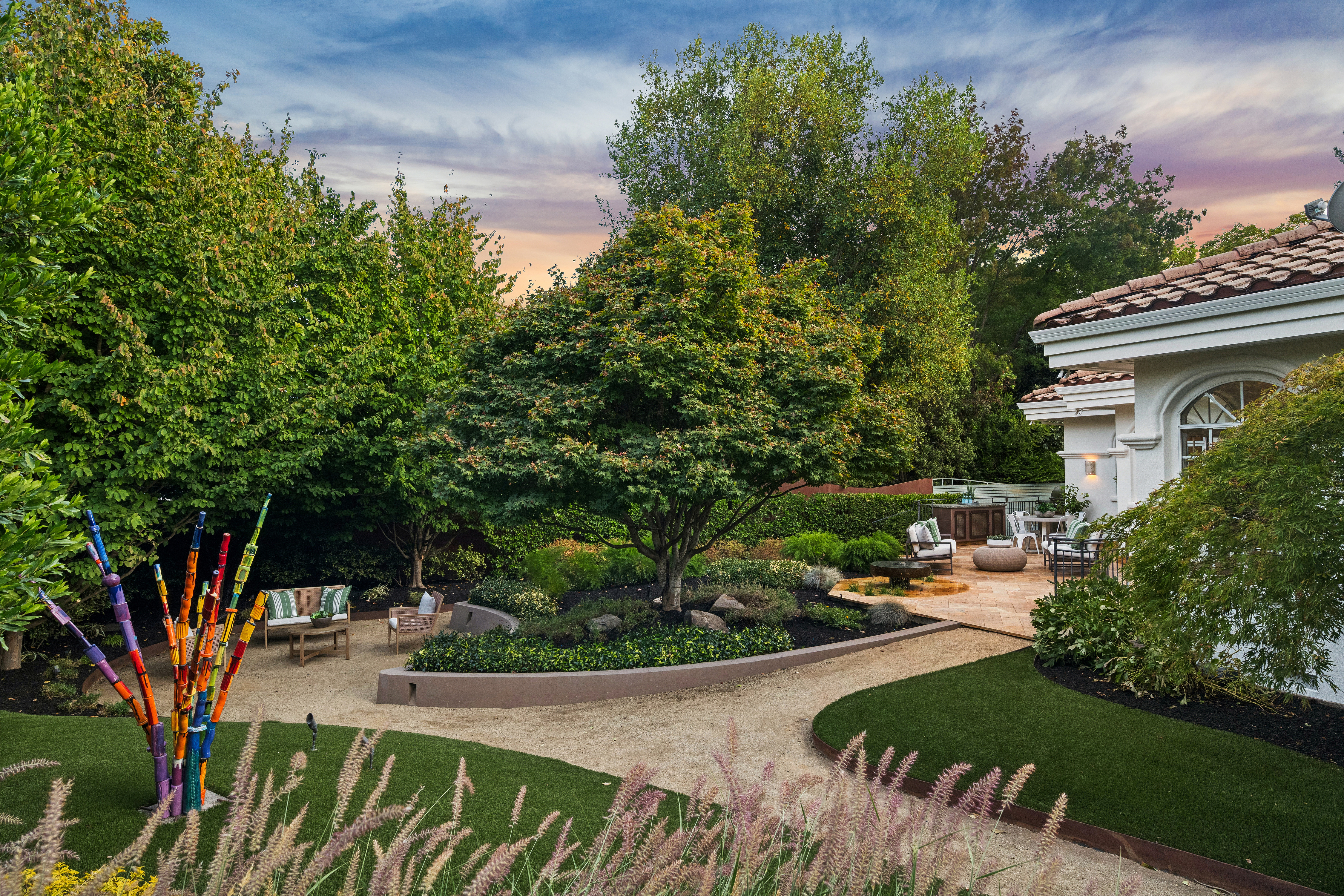 This beautifully landscaped backyard features a seamless blend of artificial turf, a flagstone patio area, and a tiered concrete retaining wall surrounding a mature shade tree. The space is accented by a vibrant, multi-colored glass art sculpture and comfortable outdoor seating areas, creating an inviting and tranquil environment for relaxation or entertaining. The overall aesthetic is elegant and meticulously maintained, highlighting a refined approach to contemporary garden design.