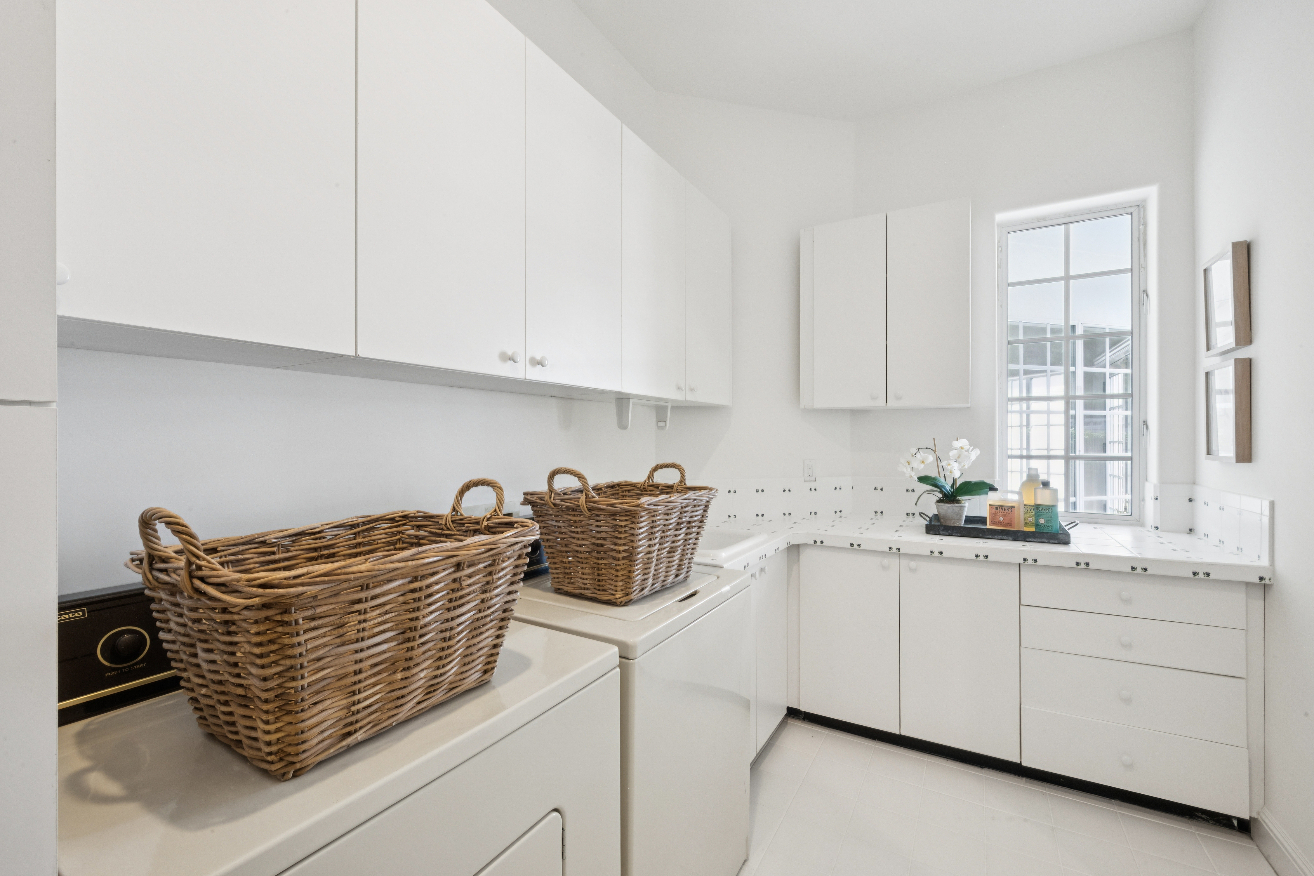 This bright and clean laundry room features white cabinetry, ample counter space, and white tiled surfaces throughout. It is equipped with a washer and dryer topped with woven baskets and includes a window that brings in natural light, making the space feel airy and functional. The room is accented with decorative items, creating a tidy and inviting atmosphere suitable for household chores.
