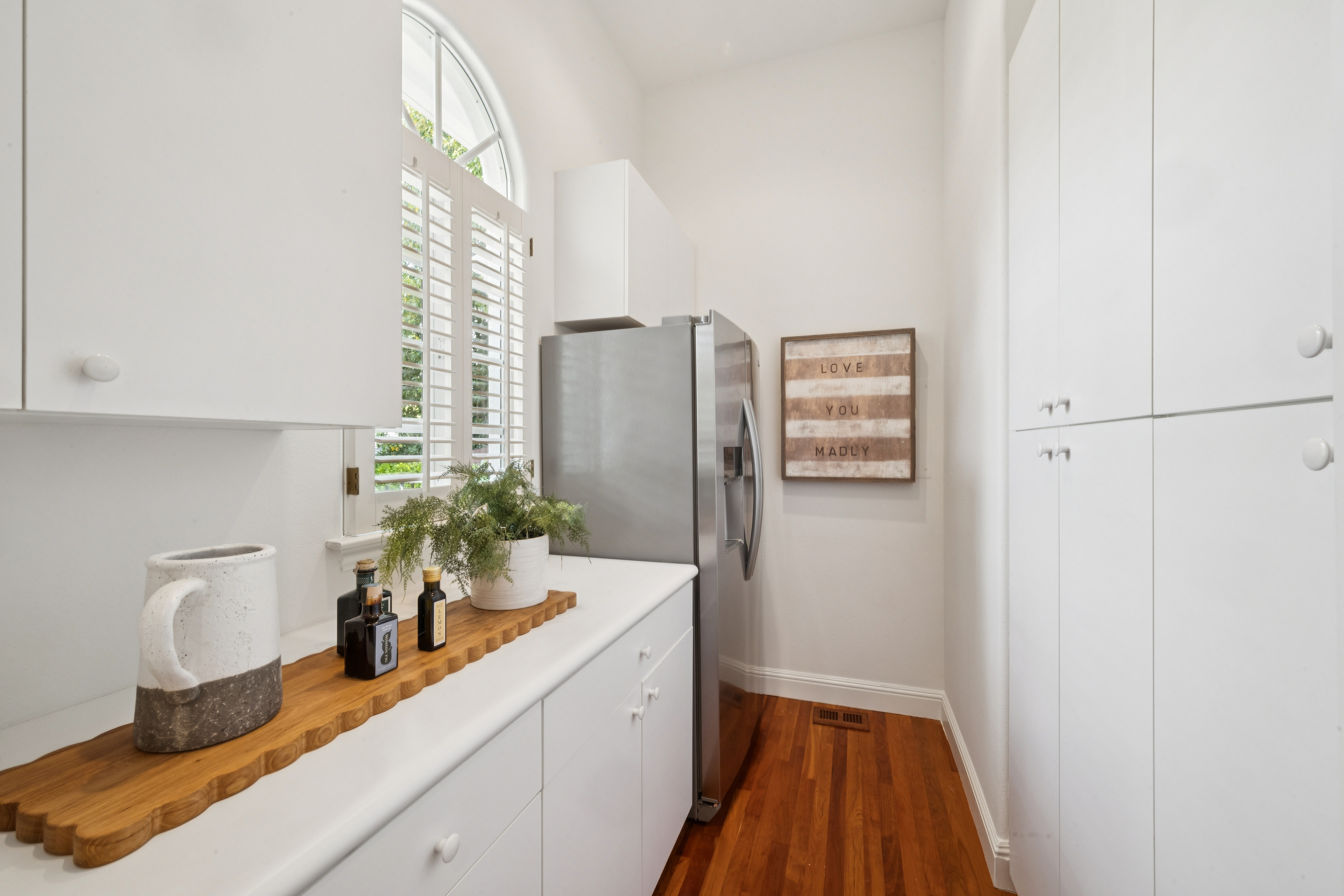 This bright and tidy kitchen area features crisp white cabinetry, stainless steel appliances, and warm hardwood flooring. A large arched window with white plantation shutters provides natural light, complemented by a decorative wooden tray and potted greenery on the countertop. A charming 'Love You Madly' wall sign adds a personalized touch to this cozy and well-maintained space.
