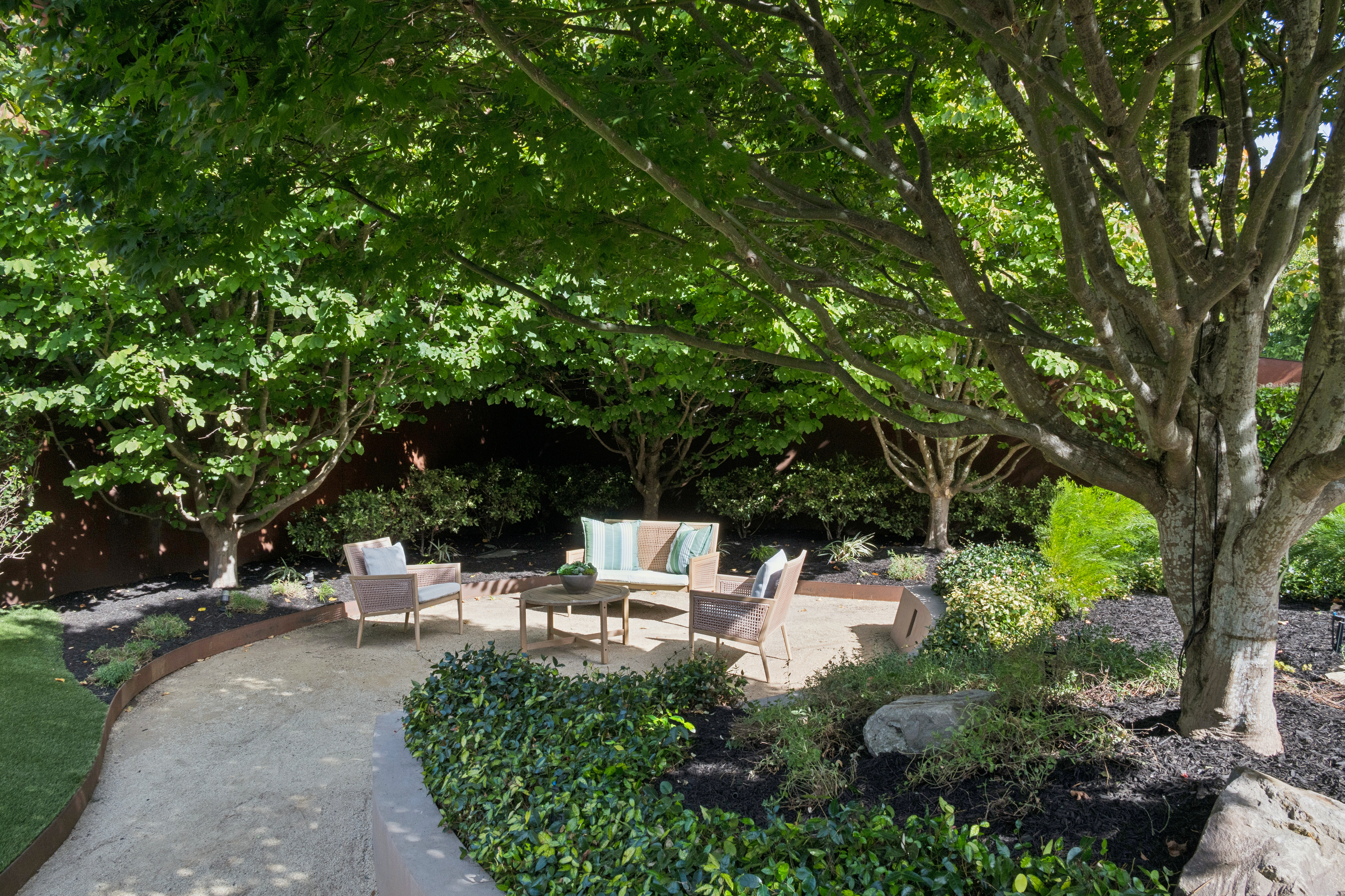 This tranquil backyard oasis features a well-defined gravel seating area nestled under a canopy of mature, lush green trees. The space includes a comfortable woven furniture set, creating an inviting spot for outdoor relaxation or entertaining. The garden is tastefully landscaped with stone borders, low-lying greenery, and mulched beds, offering a peaceful and private outdoor retreat.