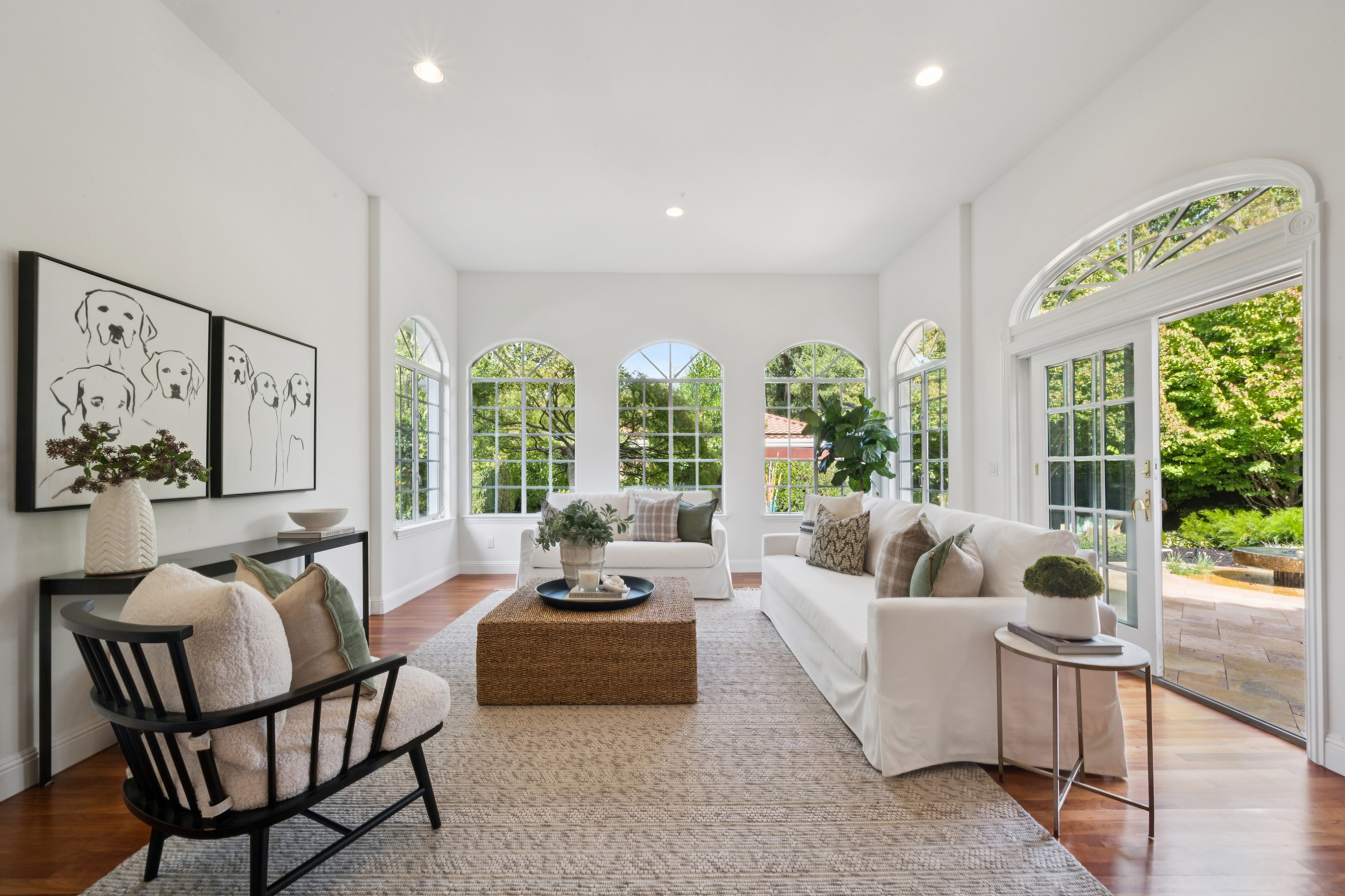 This elegant and bright living room features high ceilings, floor-to-ceiling arched windows, and French doors that provide abundant natural light and a seamless connection to the outdoor garden patio. The space is anchored by a large woven ottoman, a plush white sofa, and a modern accent chair, all set upon a neutral area rug. Its classic, sophisticated design and open, airy layout create a welcoming and serene atmosphere perfect for relaxation or entertaining.