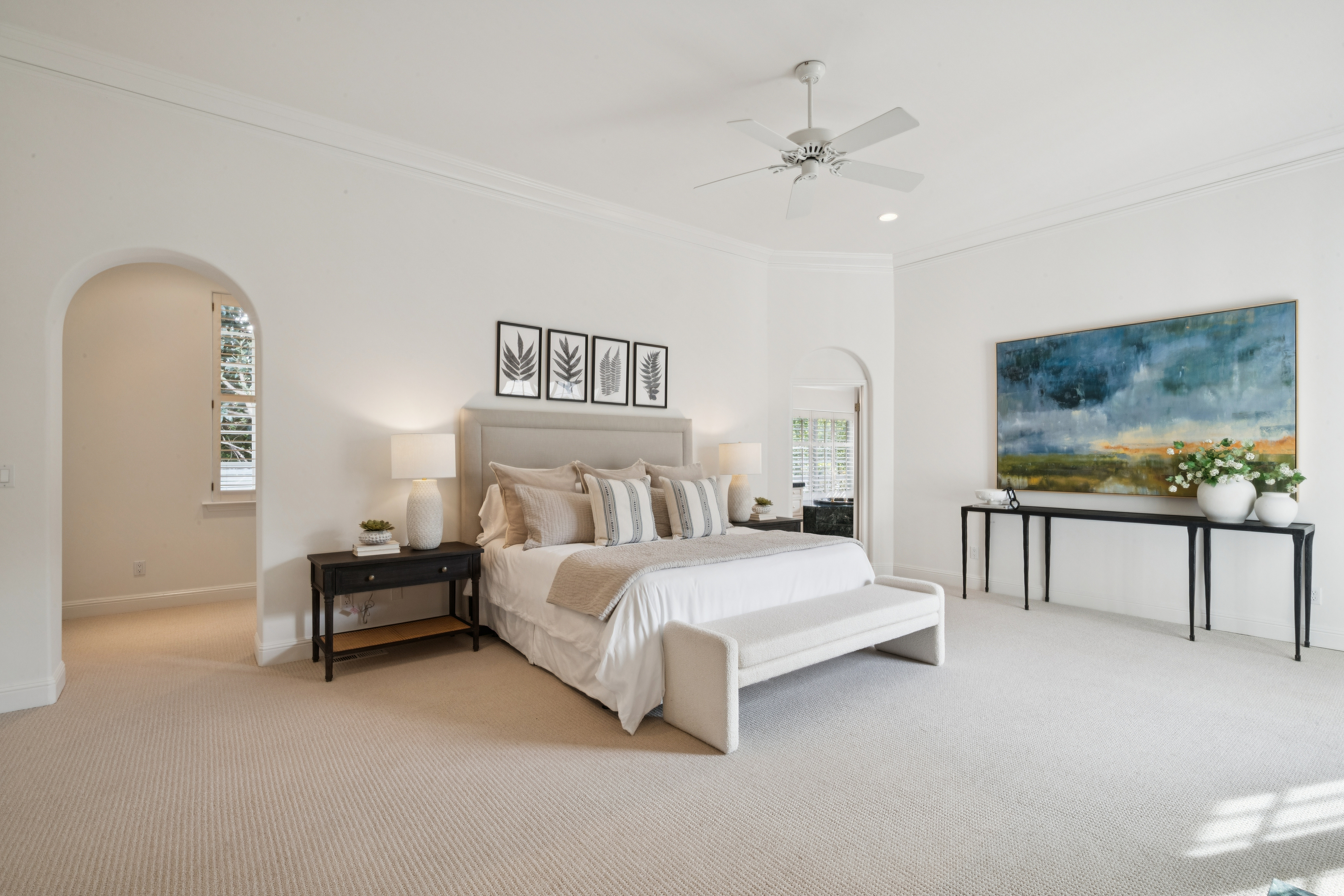 This elegant and spacious primary bedroom features a neutral color palette, creating a serene and inviting atmosphere. The room is anchored by a plush bed with an upholstered headboard, flanked by dark wood nightstands and complemented by a modern bench at the foot. A large, vibrant landscape painting hangs above a sleek console table, while arched doorways add architectural interest and a sense of refinement.