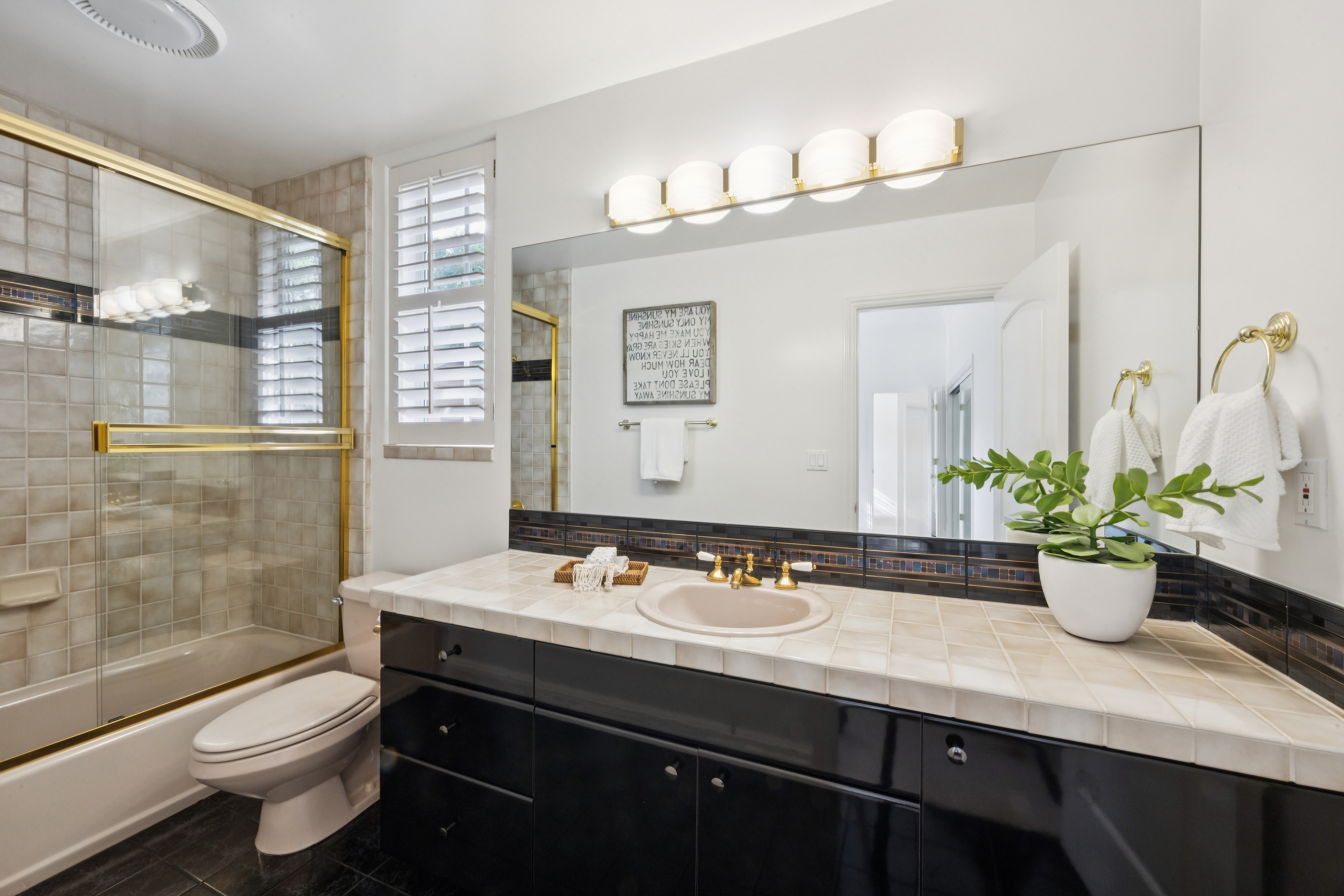 This bathroom features a bold contrast between sleek black cabinetry and light-colored tiled countertops. It includes a combined shower and tub with gold-toned hardware, a large vanity mirror reflecting the space, and a well-placed window with shutters. The combination of classic elements and modern finishes creates a clean and functional design.