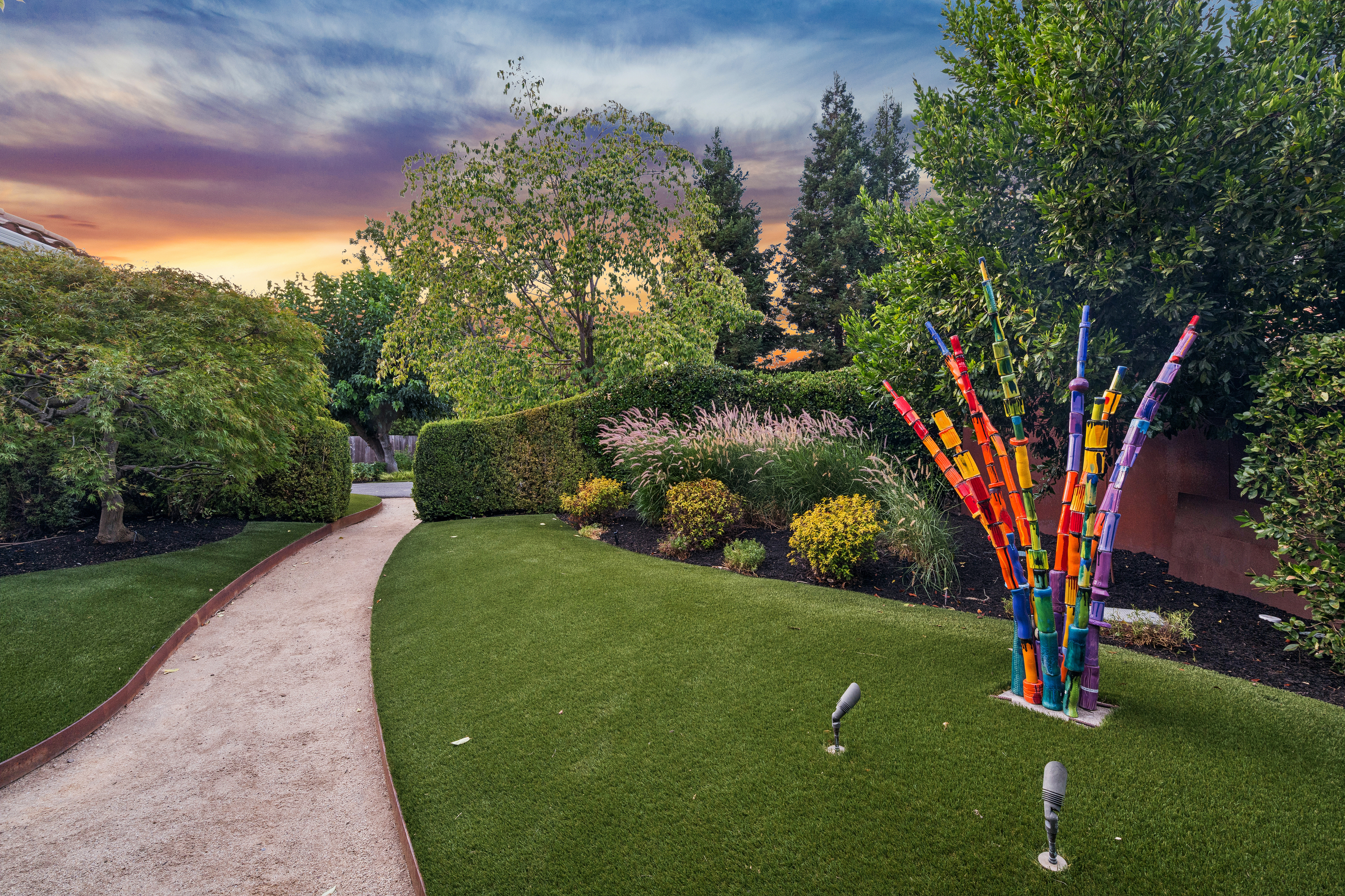 This beautifully landscaped garden features a winding gravel pathway bordered by lush green grass and manicured hedges. A vibrant, multi-colored abstract sculpture serves as a unique focal point, set against a backdrop of mature trees and a stunning sunset sky. The space offers a well-maintained, artistic outdoor aesthetic suitable for a private retreat.