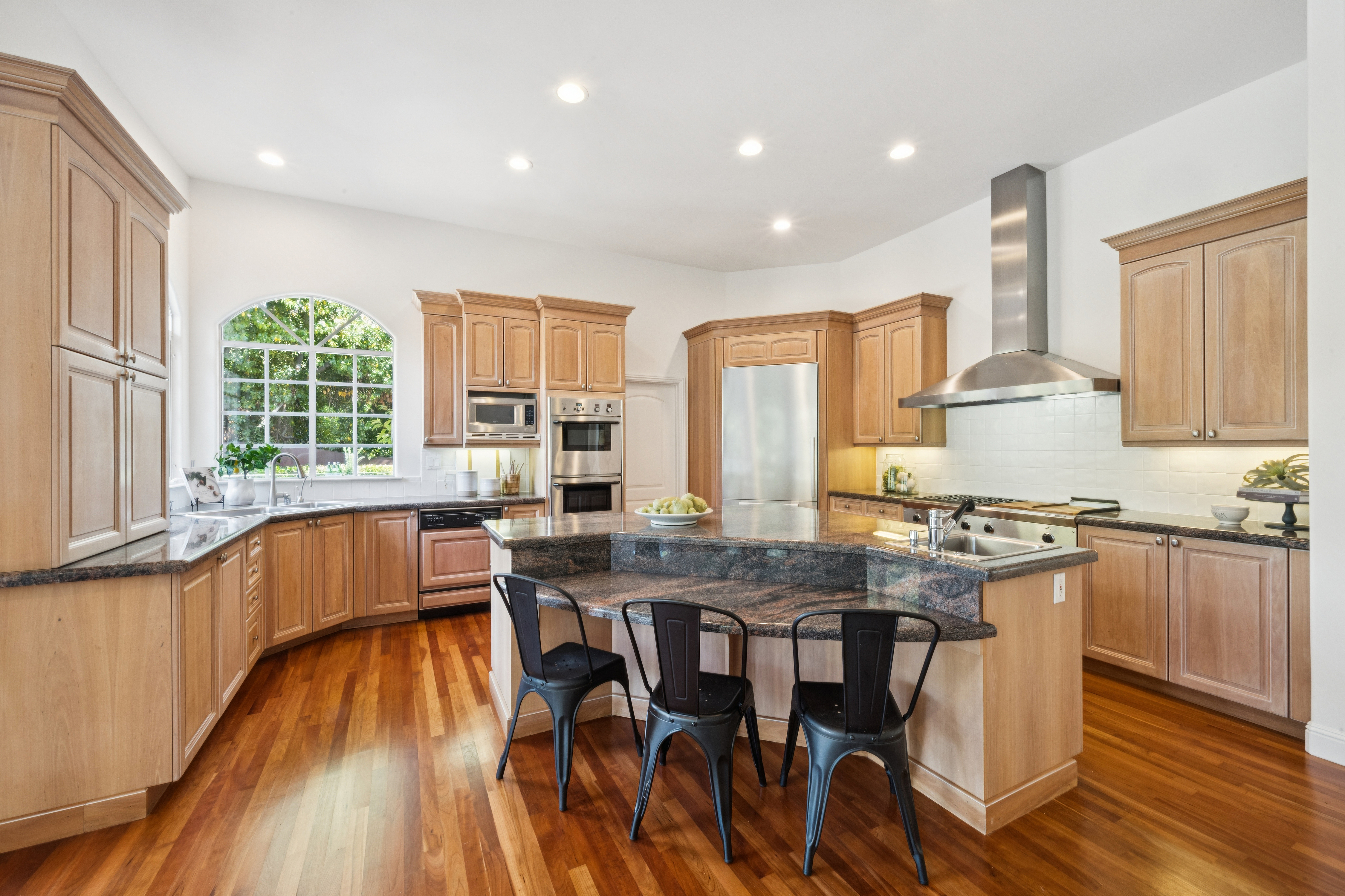 This spacious, well-lit kitchen features warm wood cabinetry, granite countertops, and high-end stainless steel appliances, including a built-in oven and microwave. The central island provides ample seating with three industrial-style metal chairs and an additional prep sink, while an arched window allows for abundant natural light. The room is completed with polished hardwood floors, creating a cohesive and inviting space perfect for cooking and entertaining.