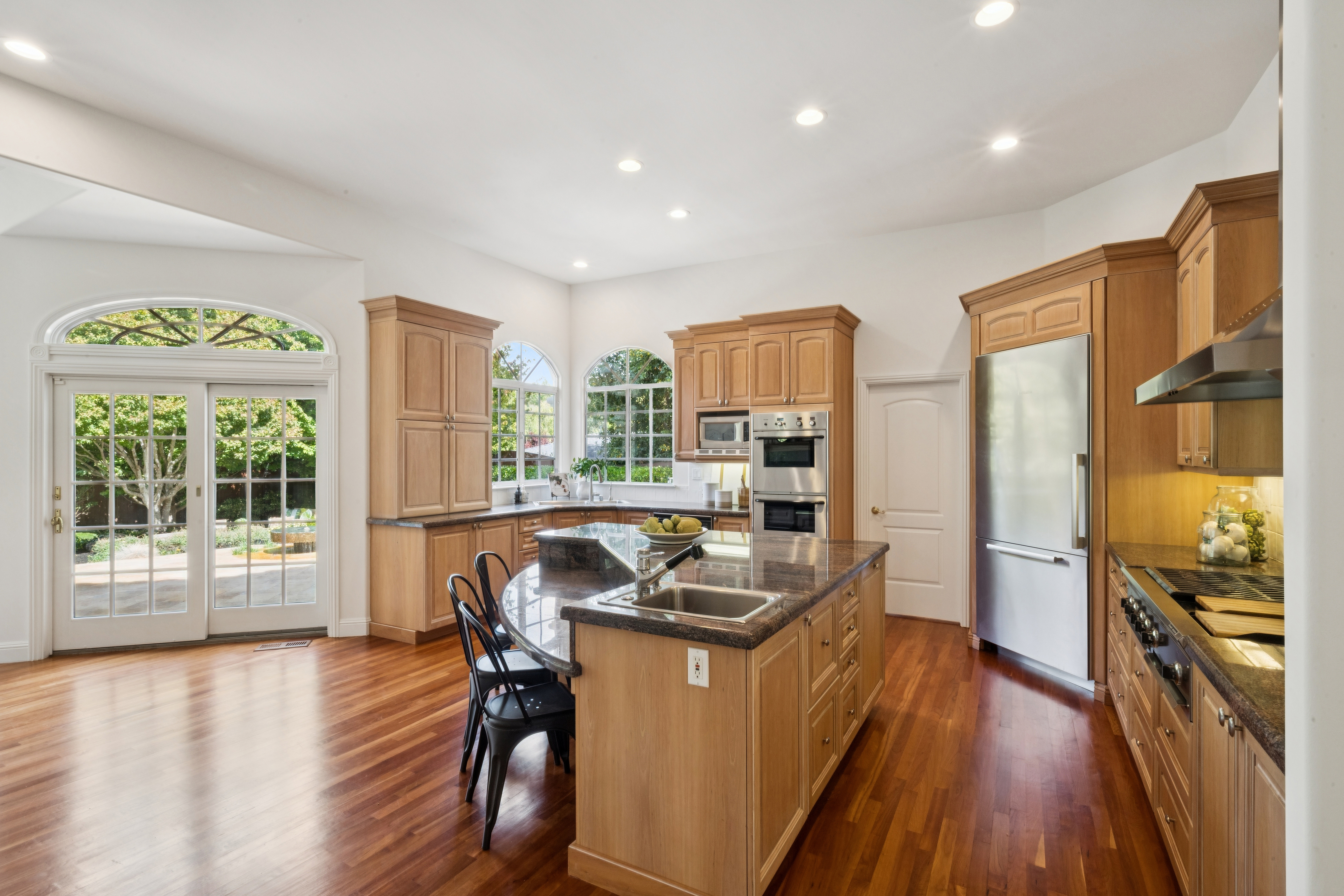 This spacious, light-filled kitchen features warm natural wood cabinetry, a large central island with granite countertops, and sleek stainless steel appliances. The open floor plan includes a dining/breakfast area connected to a patio through French doors, creating a bright and inviting atmosphere. The design is traditional and functional, enhanced by recessed lighting and a neutral palette that complements the rich hardwood flooring.
