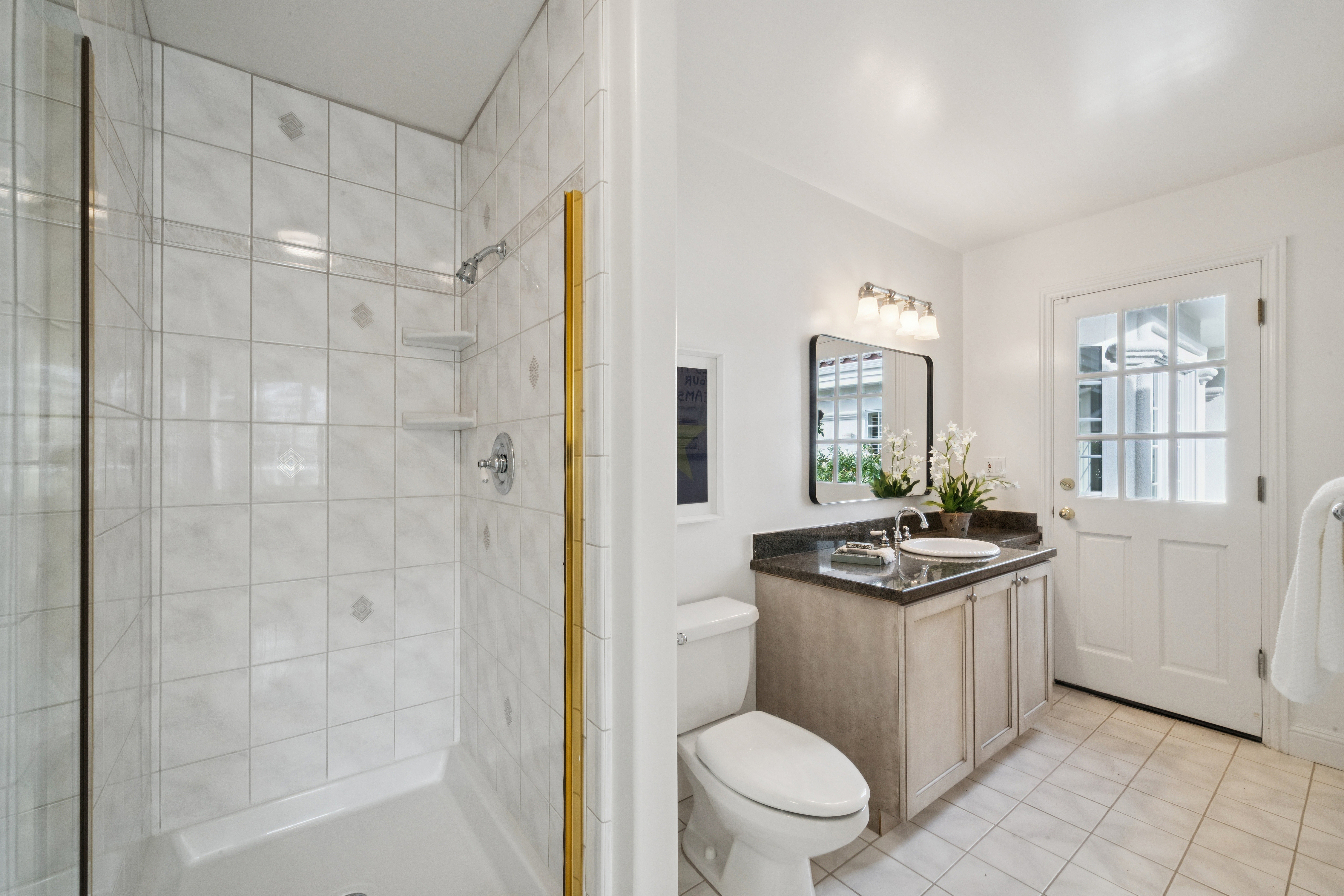 This clean and functional bathroom features a neutral color palette with light-toned wall tiles in the walk-in shower and a matching tiled floor. The vanity is topped with dark granite, complemented by a simple mirror, polished chrome fixtures, and a glass-paned door that provides direct access to the exterior. The space feels bright and well-maintained, offering a practical layout for guest use.