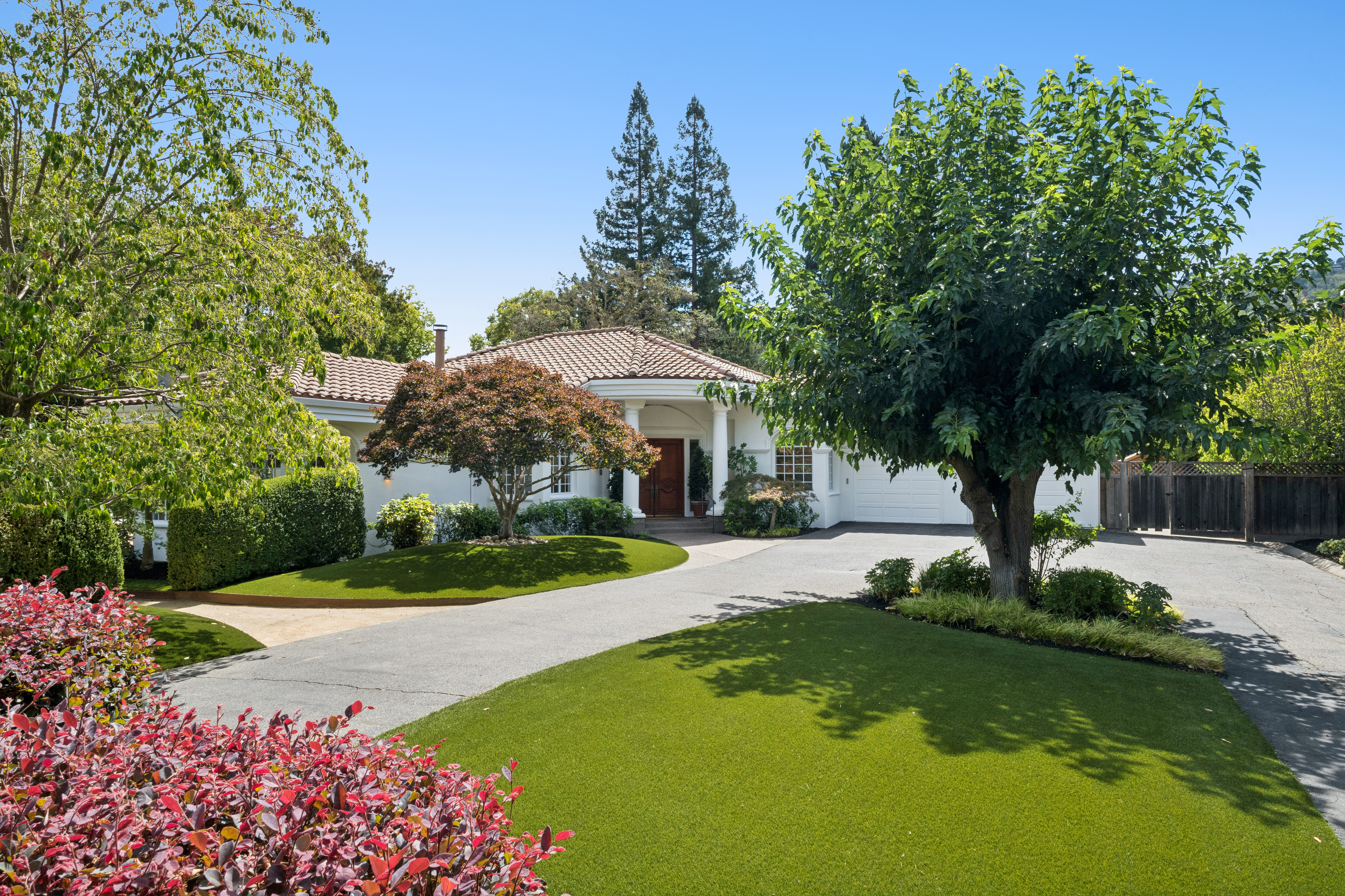 This elegant front view of a single-story home showcases a well-manicured lawn and a spacious paved driveway leading to an attached garage. The exterior features a prominent covered entryway with classic white columns and a striking wooden double door, complemented by mature trees and lush landscaping. The overall aesthetic is sophisticated and inviting, highlighting great curb appeal.