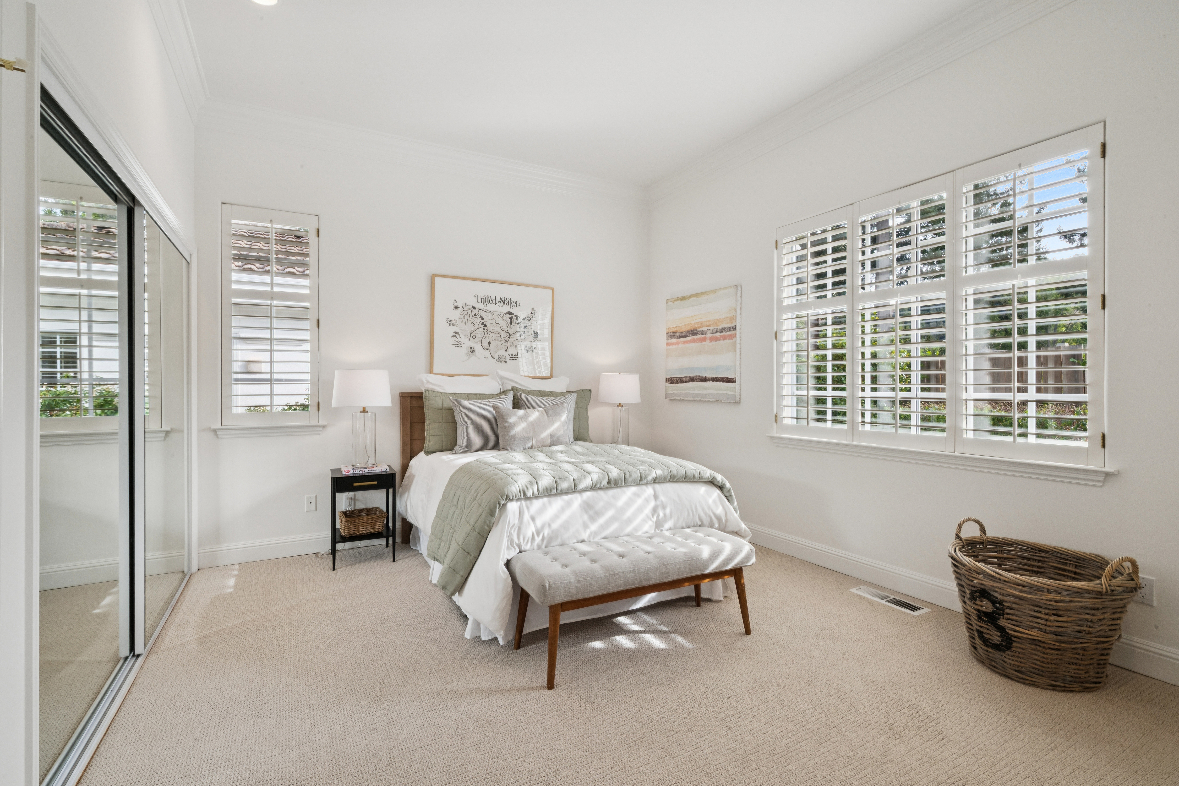 This bright and inviting bedroom features neutral tones with a cozy, layered bed, a modern tufted bench at the foot, and sleek bedside tables with minimalist lamps. The room is well-lit by windows adorned with plantation shutters, while a wall-to-wall mirrored closet adds a sense of spaciousness and reflects the clean, contemporary design.