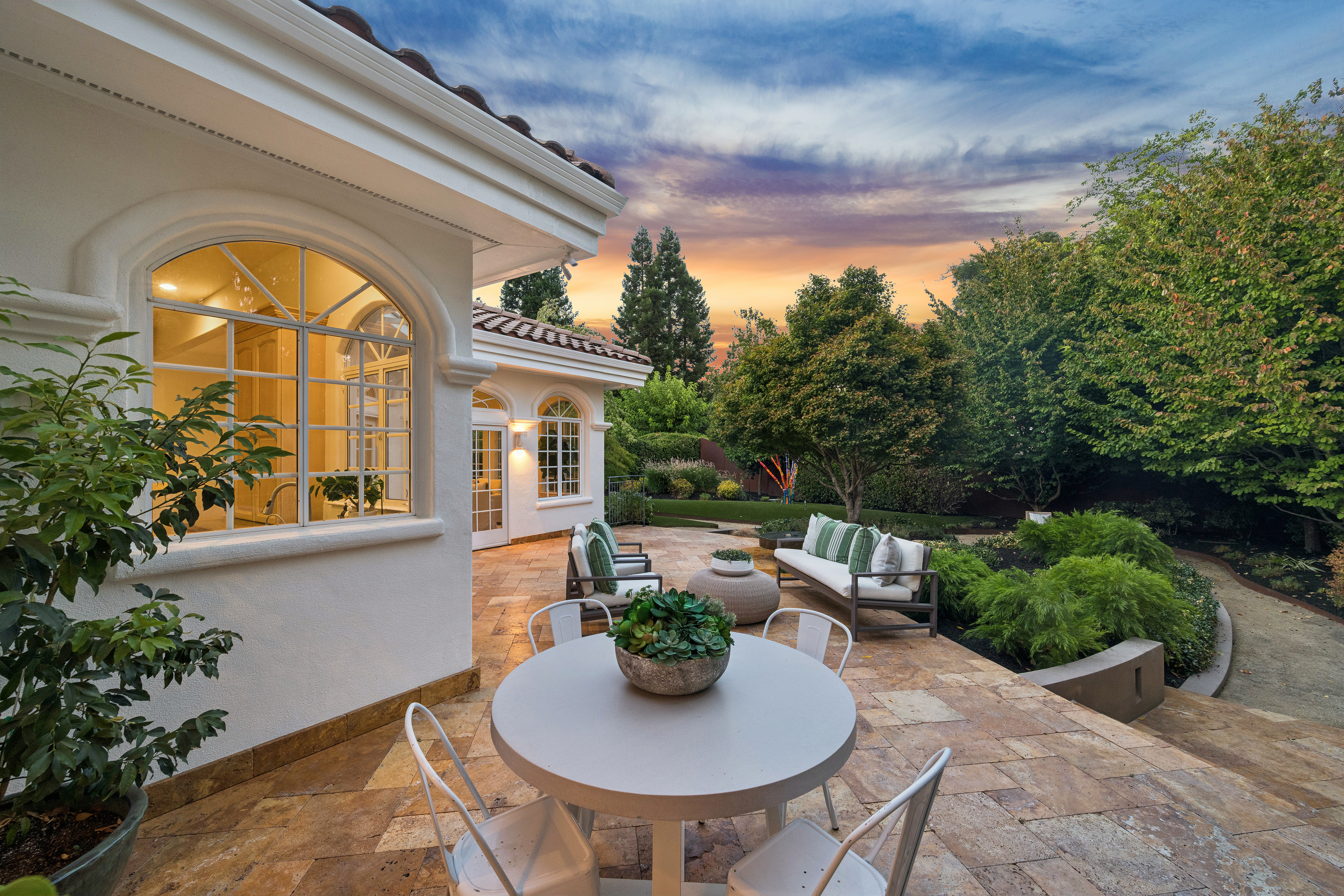 This inviting outdoor patio area features elegant stone tiling and comfortable seating arrangements, including a round bistro set and a modern sofa. The space is set against a clean white-stucco home with classic arched windows, looking out onto a lush, manicured backyard at sunset. It provides a serene and well-maintained setting perfect for outdoor relaxation or hosting gatherings.