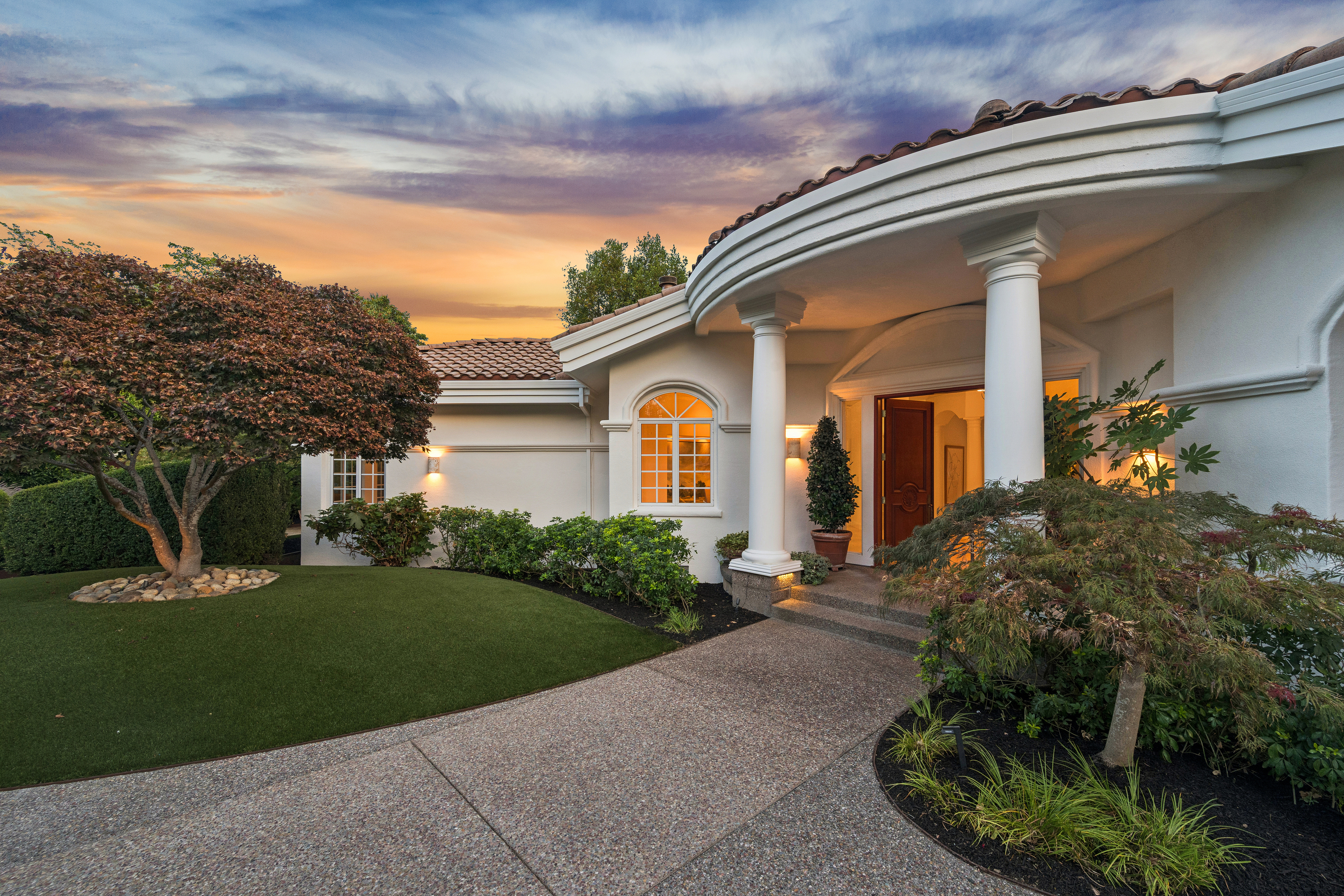 This inviting home exterior features a graceful, curved entry portico supported by classic white columns. The scene highlights a well-manicured lawn, mature landscaping, and a textured concrete walkway leading to a stately front door, all set against a stunning sunset sky. The architecture exudes a timeless, elegant curb appeal that makes for a welcoming entryway.