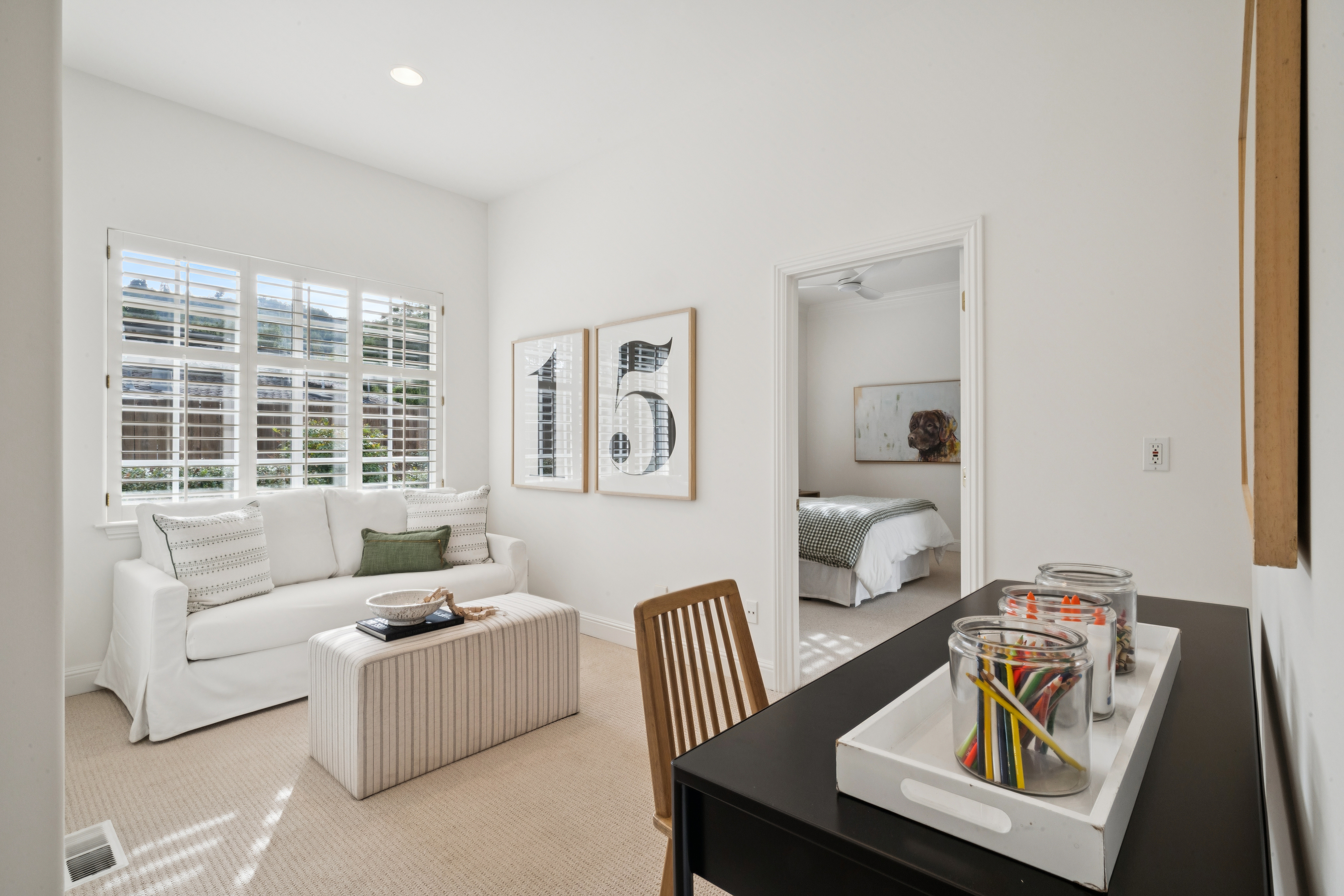 This bright and inviting multi-functional room serves as a comfortable den or office, featuring a clean white sofa, a striped ottoman, and a practical workspace area. The neutral color palette and soft natural light streaming through the shuttered windows create a serene and flexible environment. A doorway provides an appealing view into an adjacent bedroom, enhancing the home's sense of flow and cohesive design.