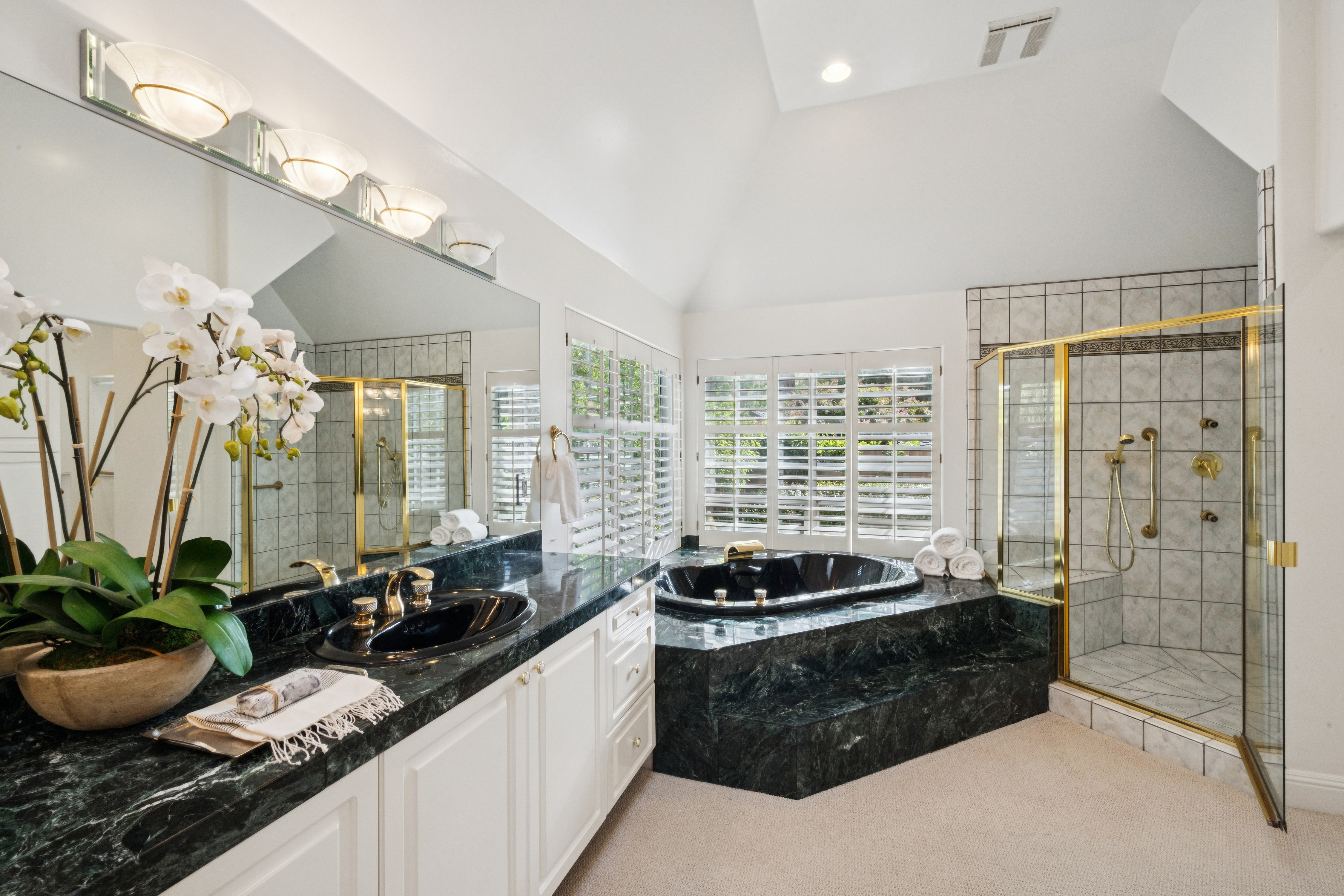 This luxurious primary bathroom features a bold design with deep green marble countertops and matching built-in steps for the large, integrated soaking tub. The space includes a glass-enclosed shower with gold fixtures, a dual-sink vanity with a large wall mirror, and classic white plantation shutters. High ceilings and tasteful decorative accents like a potted orchid contribute to an elegant, spa-like atmosphere.