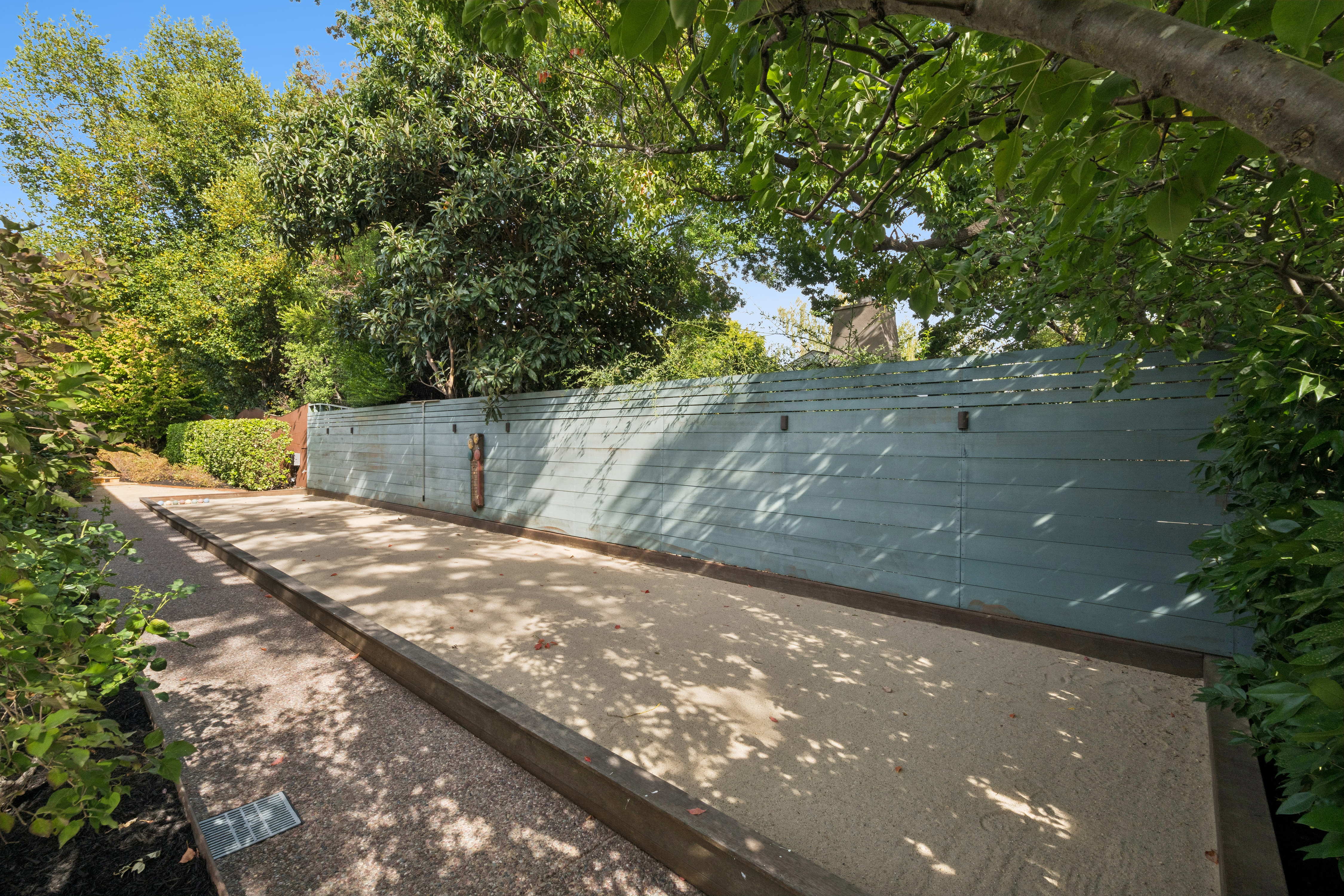 This outdoor space features a well-maintained bocce ball court set within a serene, lush garden environment. The area is enclosed by a stylish horizontal slat fence painted in a soft blue-grey, providing both privacy and a contemporary aesthetic. With surrounding mature trees and manicured foliage, this amenity offers an inviting and recreational setting for outdoor leisure.