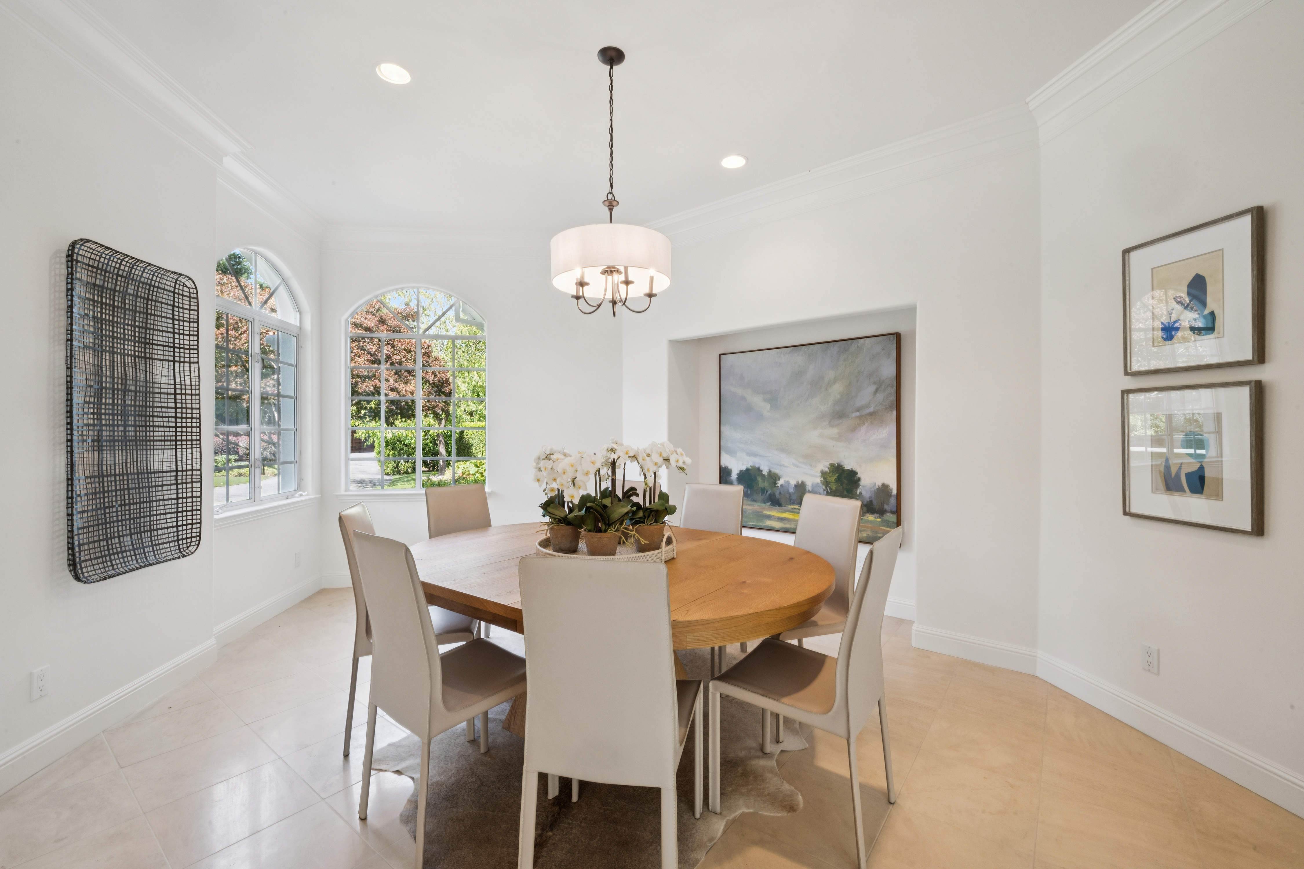 This elegant dining room features a round wooden table surrounded by sleek, beige upholstered chairs, creating a sophisticated and inviting space. The room is accented with a central drum pendant light, a large landscape painting set into a niche, and two arched windows that flood the area with natural light. The neutral color palette and clean lines provide a timeless and polished look perfect for formal entertaining.