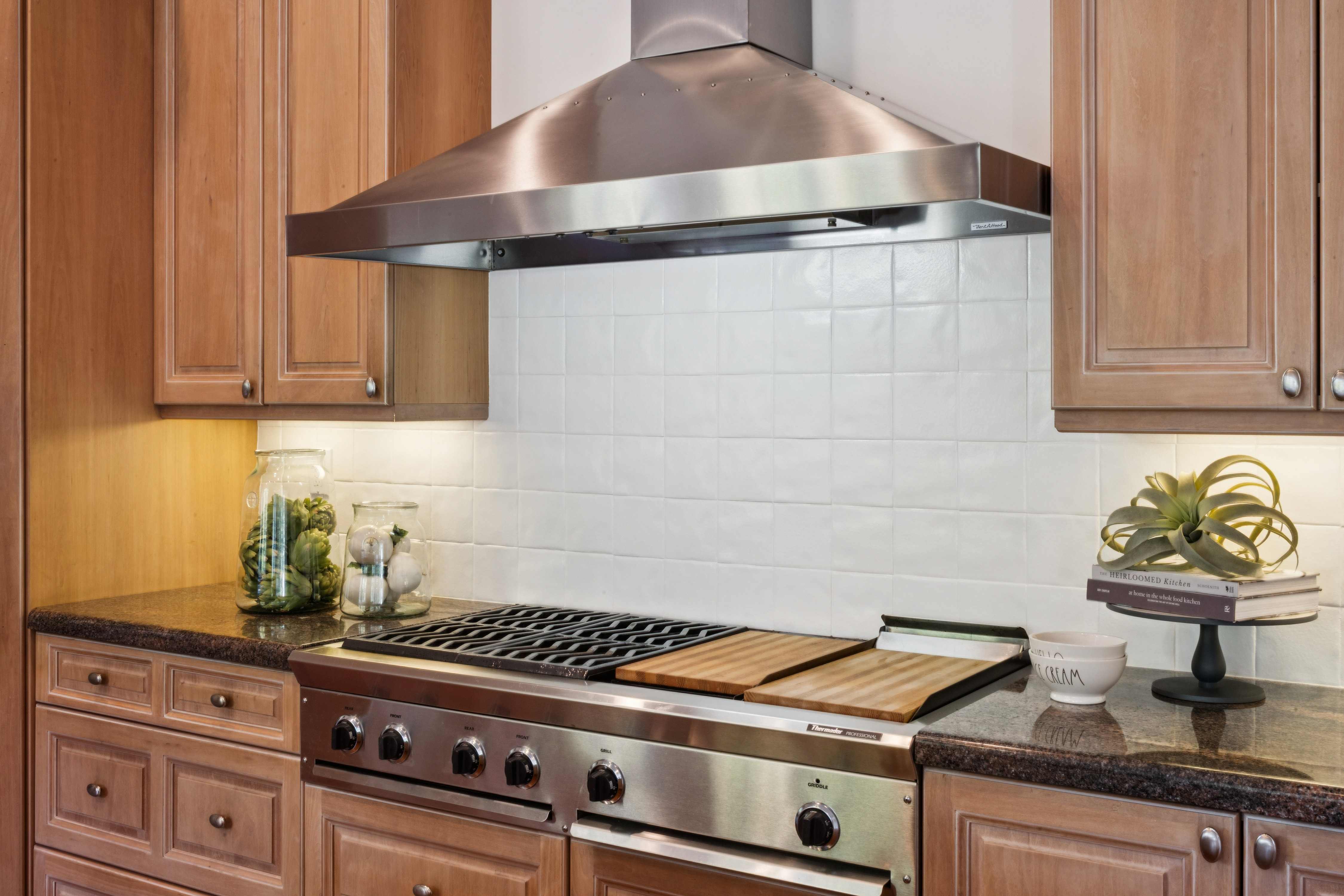 This kitchen area features a high-end stainless steel Thermador professional range equipped with a griddle and wooden cutting board inserts. The space is anchored by a matching stainless steel range hood and a classic white square-tile backsplash, complemented by traditional warm-toned wood cabinetry and dark granite countertops. This professional-grade setup offers a sophisticated and functional aesthetic perfect for a gourmet home.