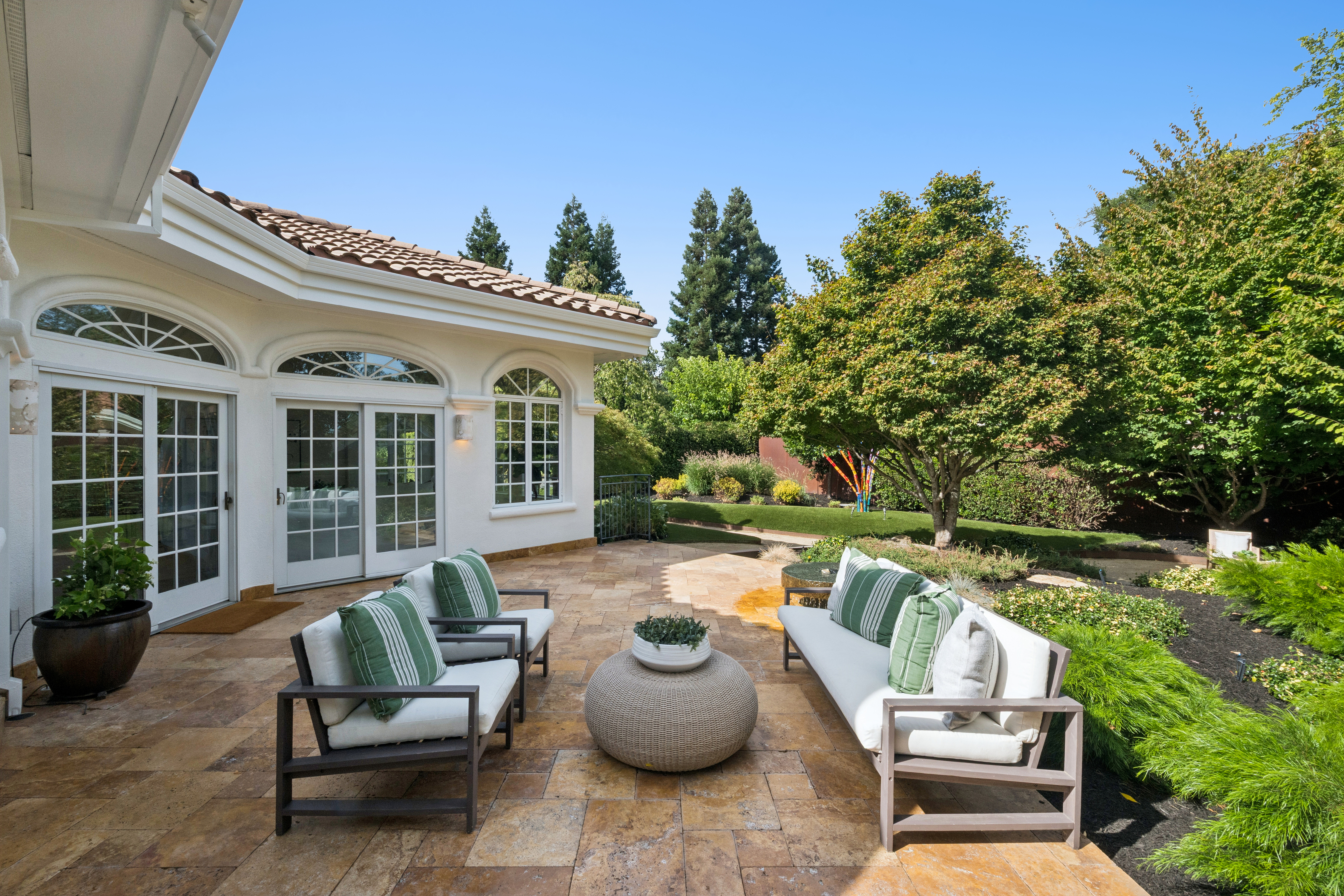This inviting outdoor living space features a spacious stone-tiled patio furnished with modern cushioned seating, including two armchairs and a matching sofa around a circular woven coffee table. The area is seamlessly integrated with the home's white stucco exterior, which includes elegant arched windows and sliding glass doors that open to the patio. Surrounded by lush greenery, a manicured lawn, and mature trees, this tranquil setting offers a perfect retreat for relaxation or entertaining in a private garden-like environment.