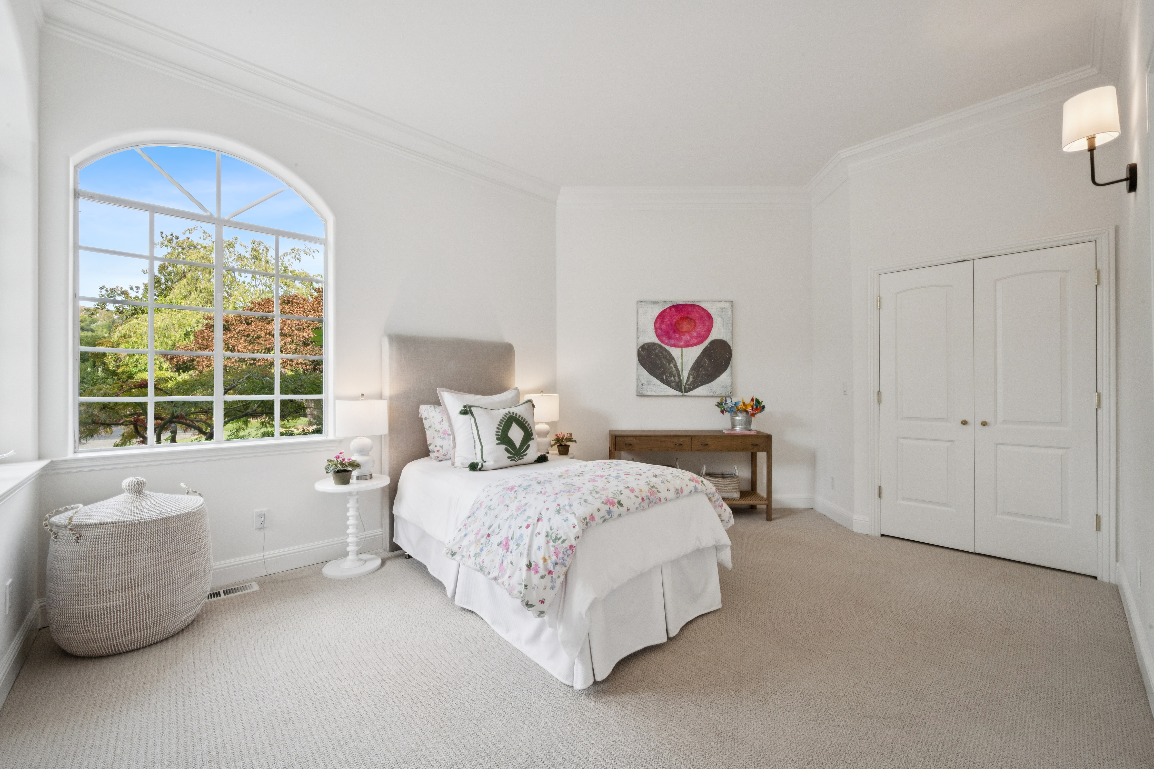This bright and inviting bedroom features a twin-sized bed with a floral quilt, complemented by a large, arched window that fills the space with natural light. The room is decorated with a clean, neutral palette and includes a wooden console table, a small side table, and a decorative wicker basket. A wall-mounted sconce and elegant crown molding complete the polished, comfortable aesthetic.
