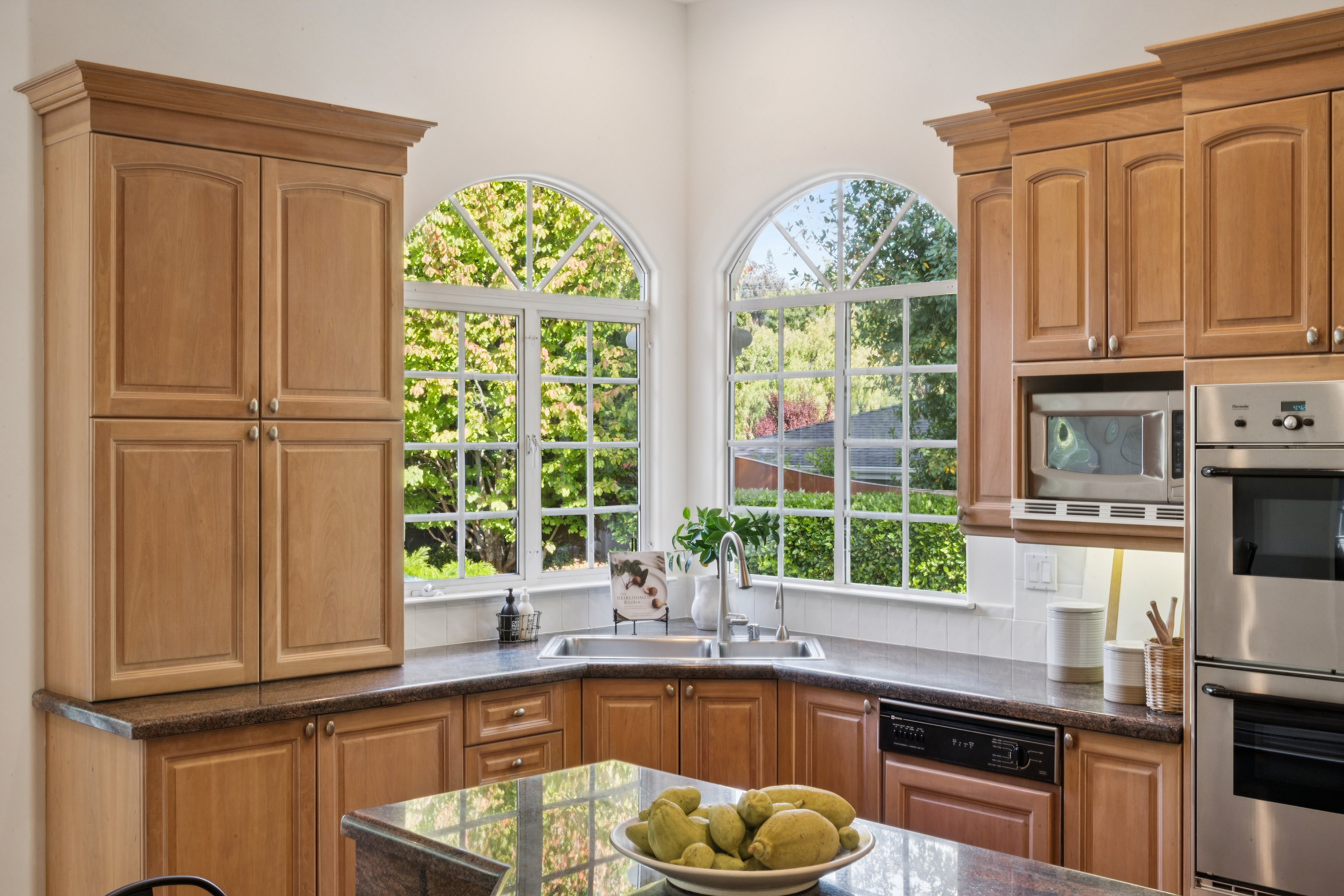 This kitchen features warm-toned wooden cabinetry and dark granite countertops, creating a traditional and inviting aesthetic. A corner sink setup sits beneath two large, arched windows that provide natural light and a pleasant view of the outdoor greenery. Equipped with stainless steel built-in ovens and a microwave, the space is functional and well-organized for culinary activities.