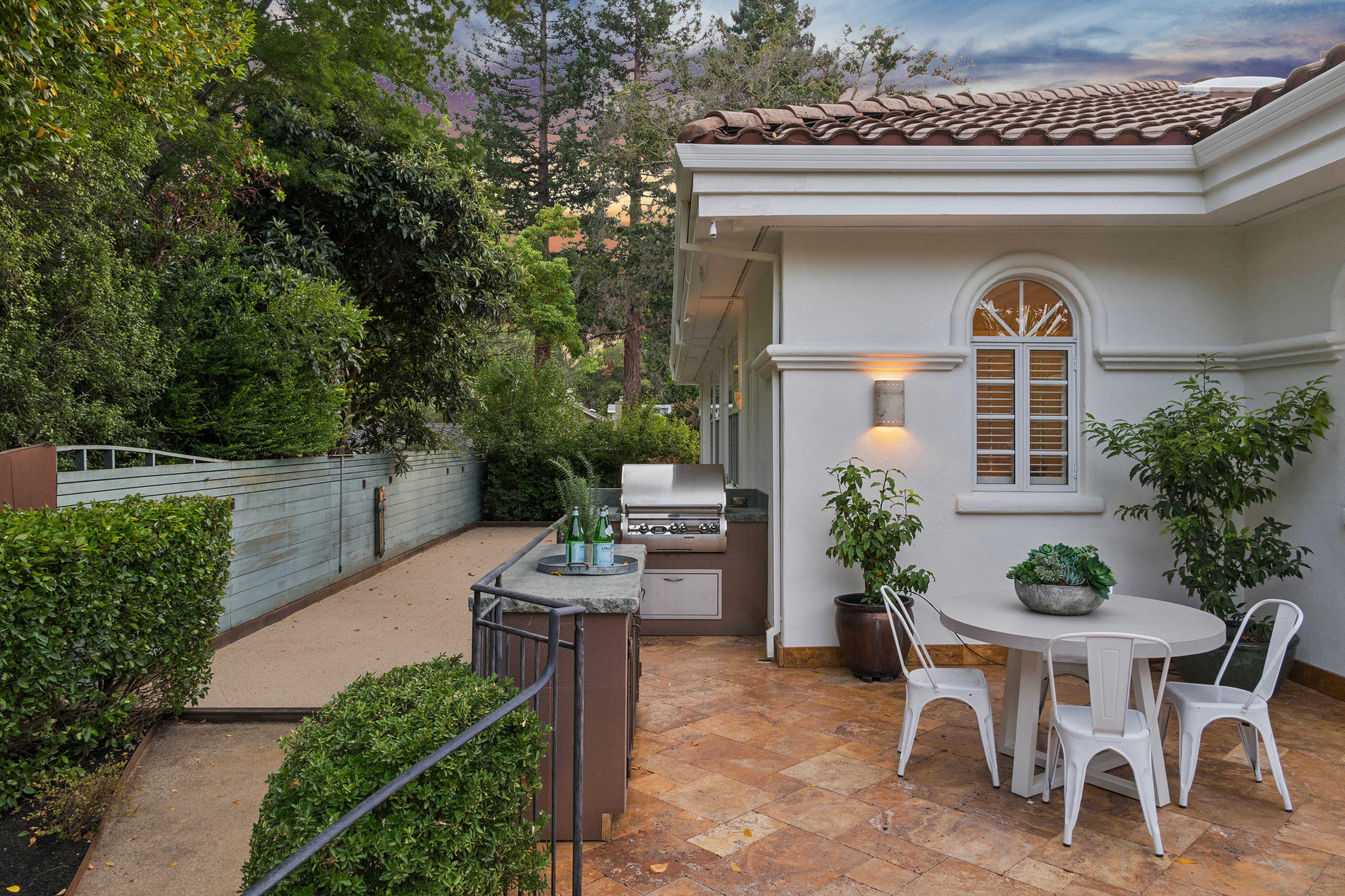 This outdoor patio area features a sophisticated built-in stainless steel grill and stone-topped island, perfect for al fresco cooking and dining. The space is accented by a charming round table with white metal seating, surrounded by lush potted plants and elegant architectural details like the arched window. Its warm, tiled flooring and intimate atmosphere create an inviting extension of the home's living space.